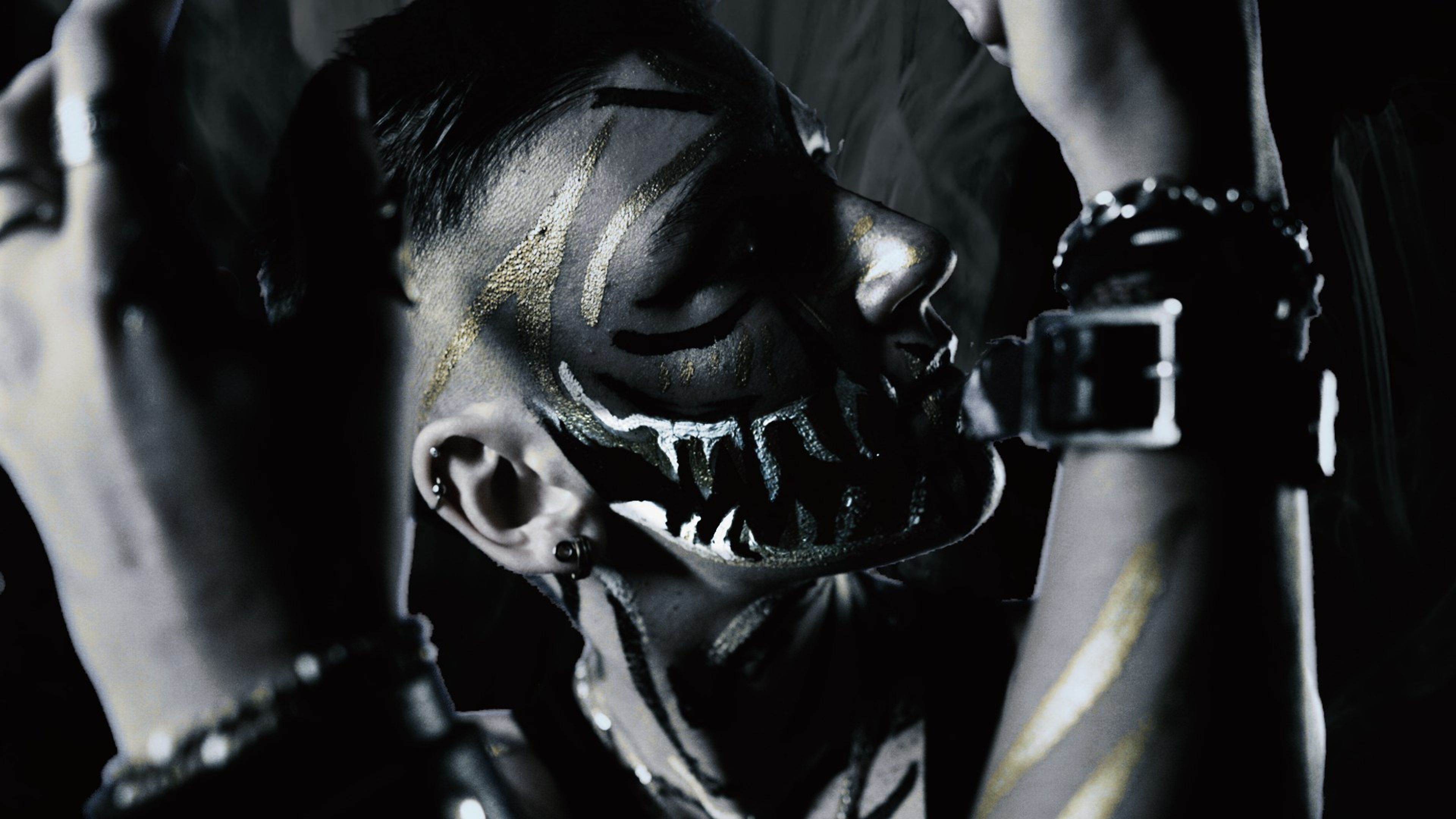 Close profile of a face painted with black fangs and gold lines, hands and a studded wristband framing it