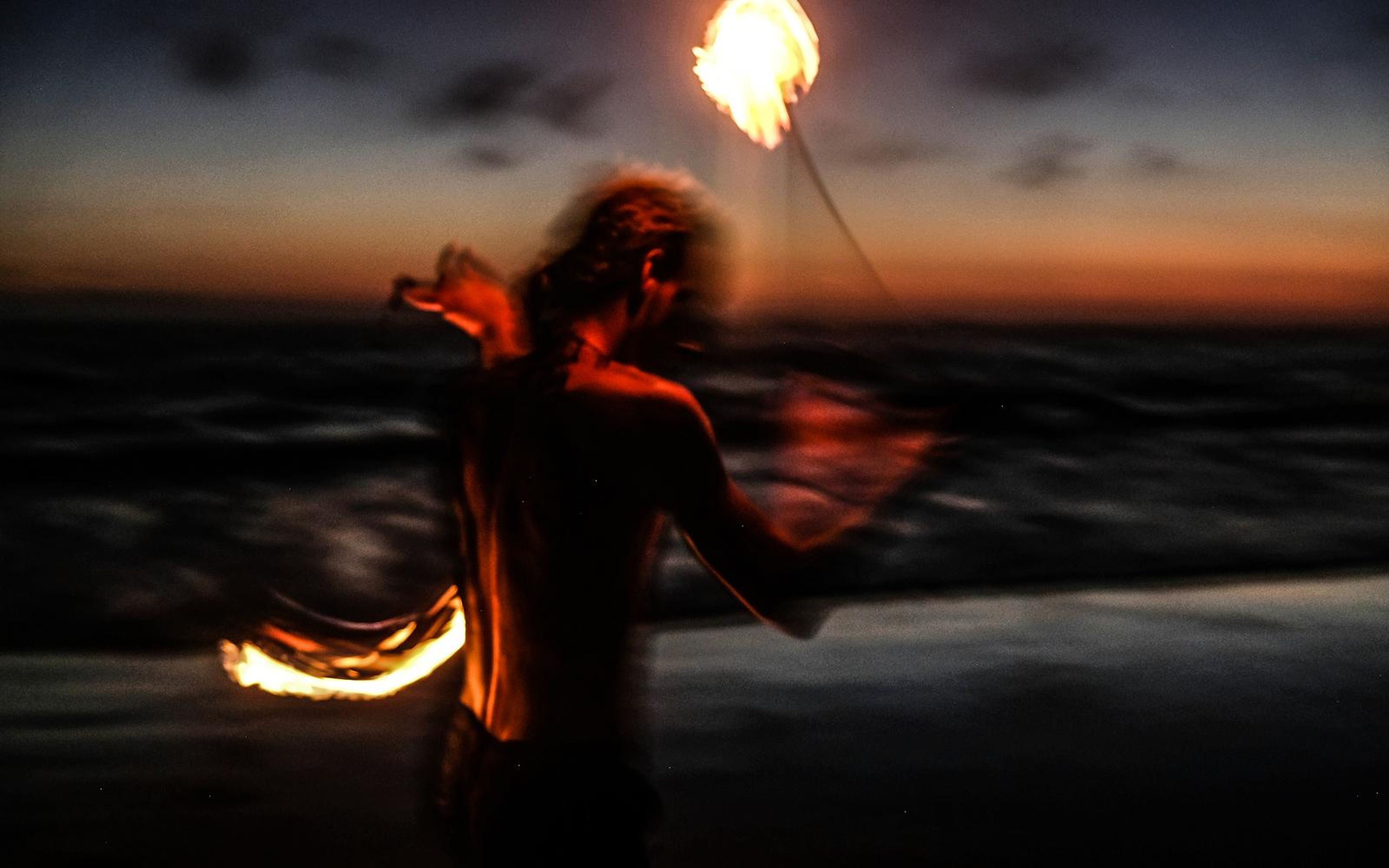 Motion-blurred performer on the shore swinging fire poi, with sunset glow and waves behind