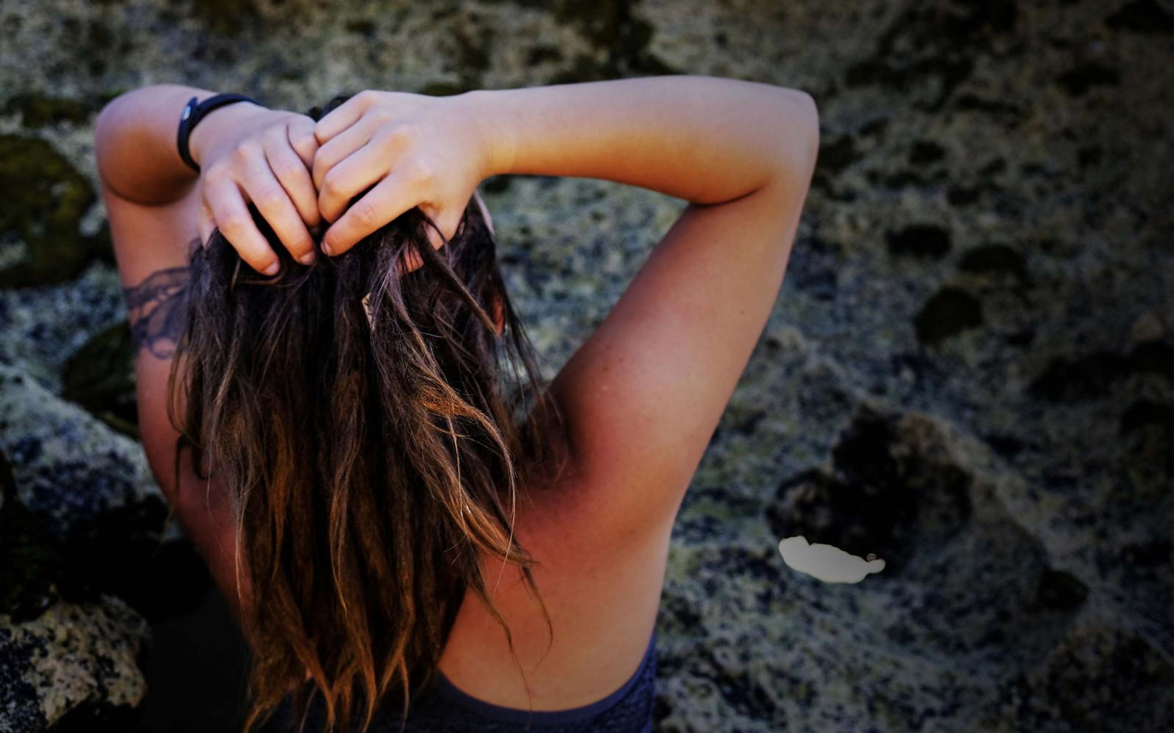 Woman seen from behind with both hands in her long hair, dark lichen-covered rock behind