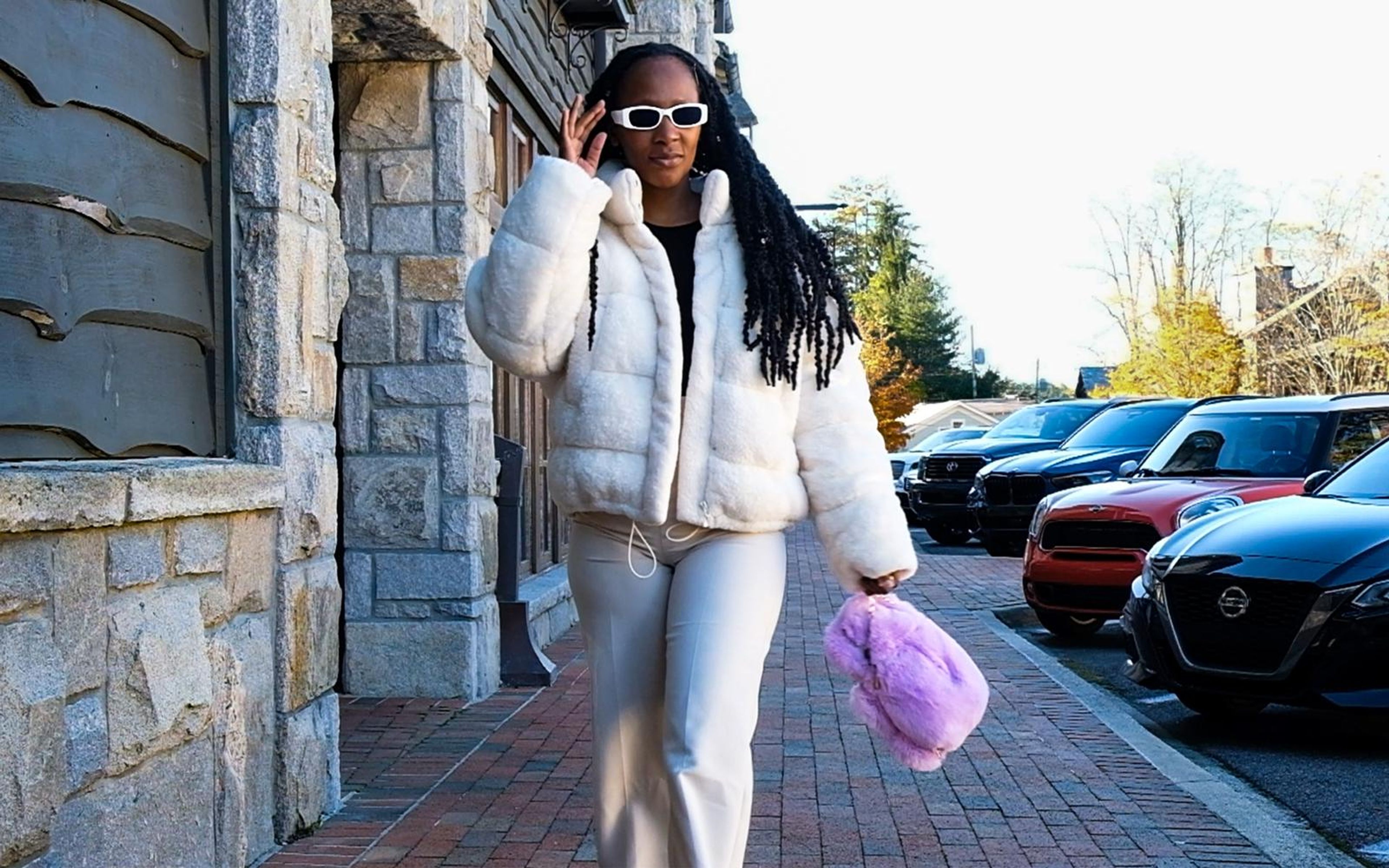 Model walking along a brick pavement, hand on white sunglasses, white fur jacket, purple fluffy bag