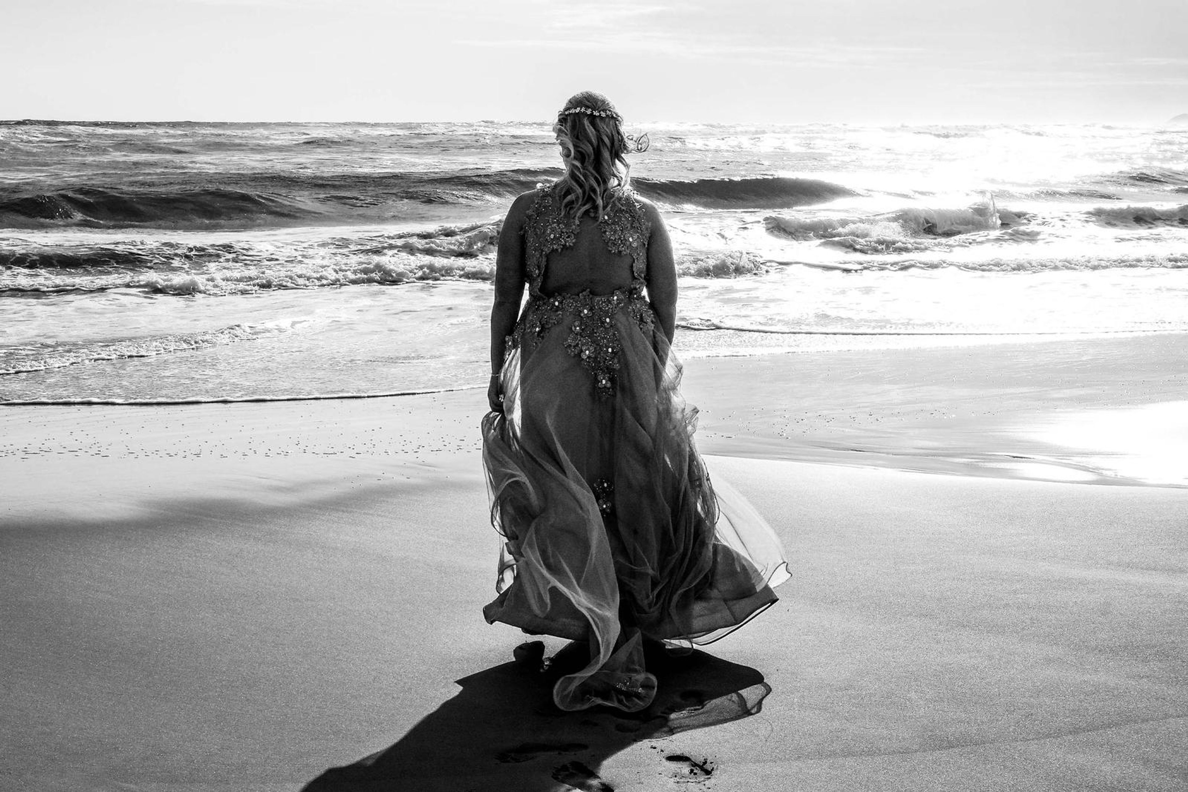Black and white back view with the gown trailing across the wet sand