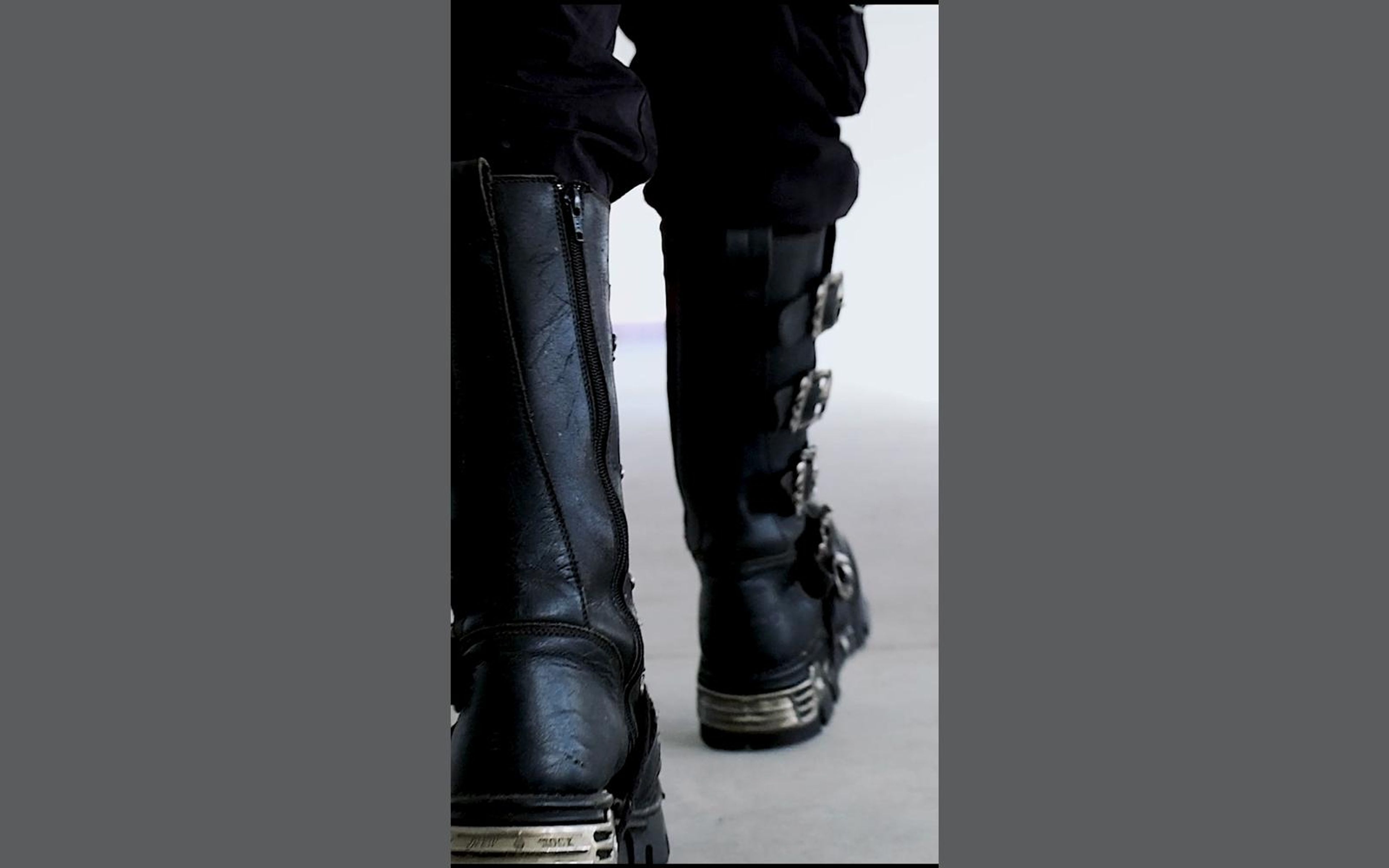 Low view of a laced, buckled New Rock boot planted on grey concrete beside the other leg