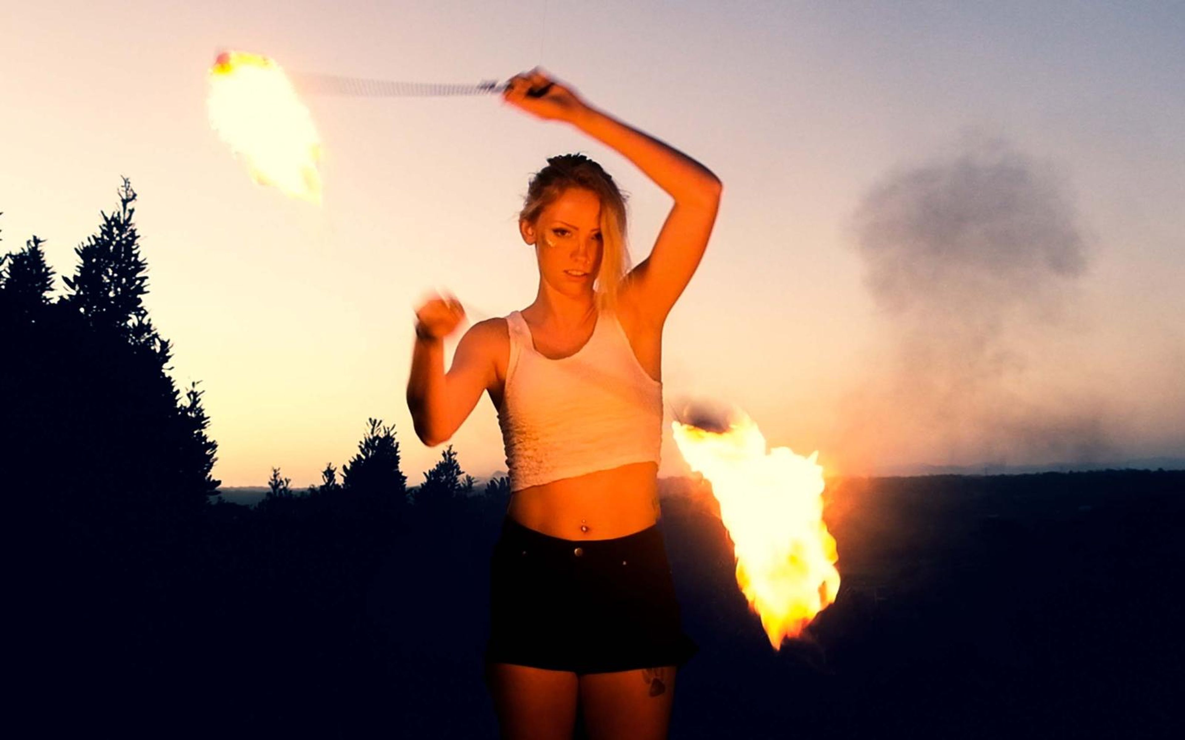 Performer in a white crop top and dark shorts spinning fire poi, smoke at right and trees silhouetted at dusk