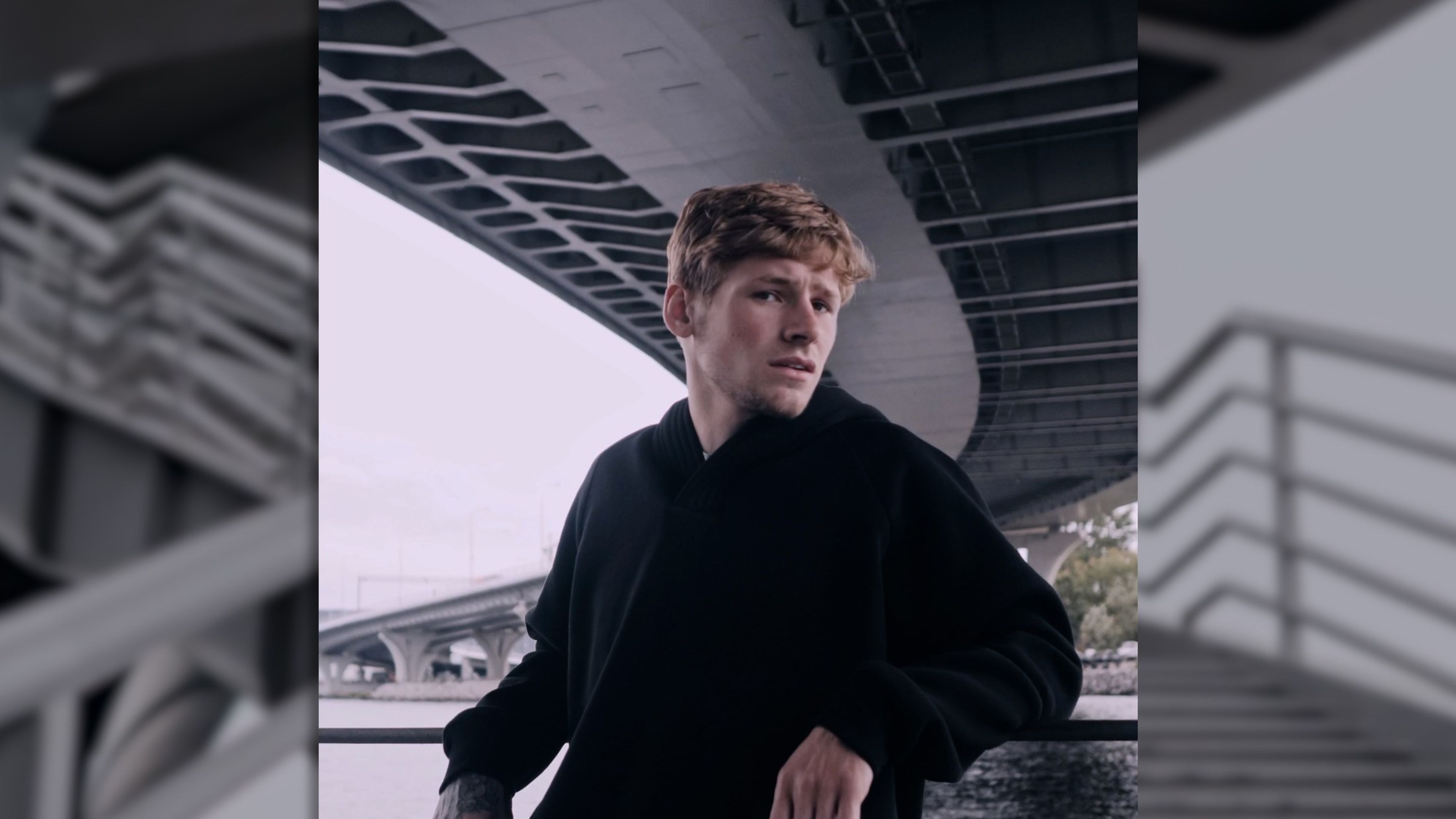 Man with ginger hair in a black top posed beneath a concrete overpass