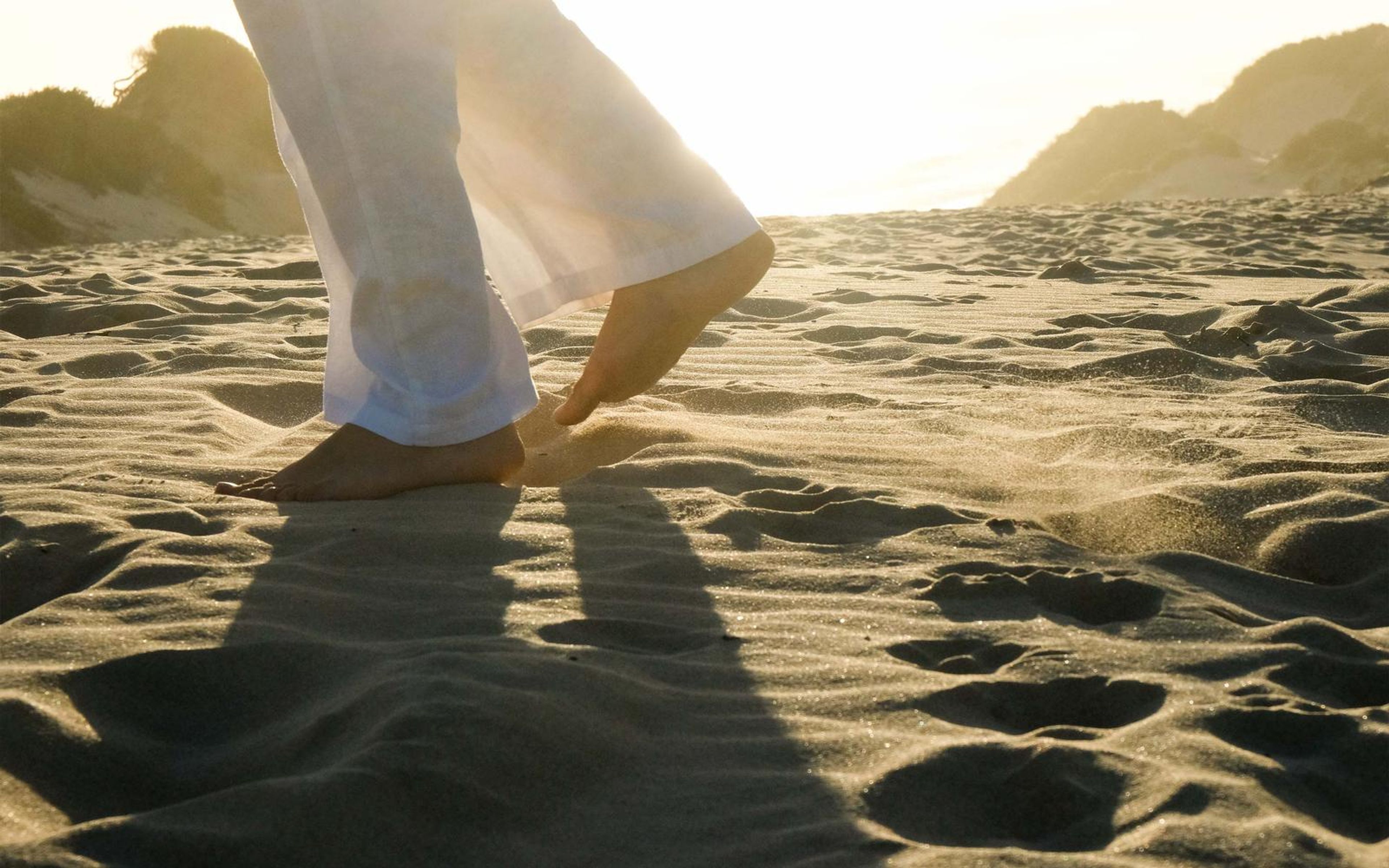 Bare feet and white trousers walking across rippled sand with long shadows