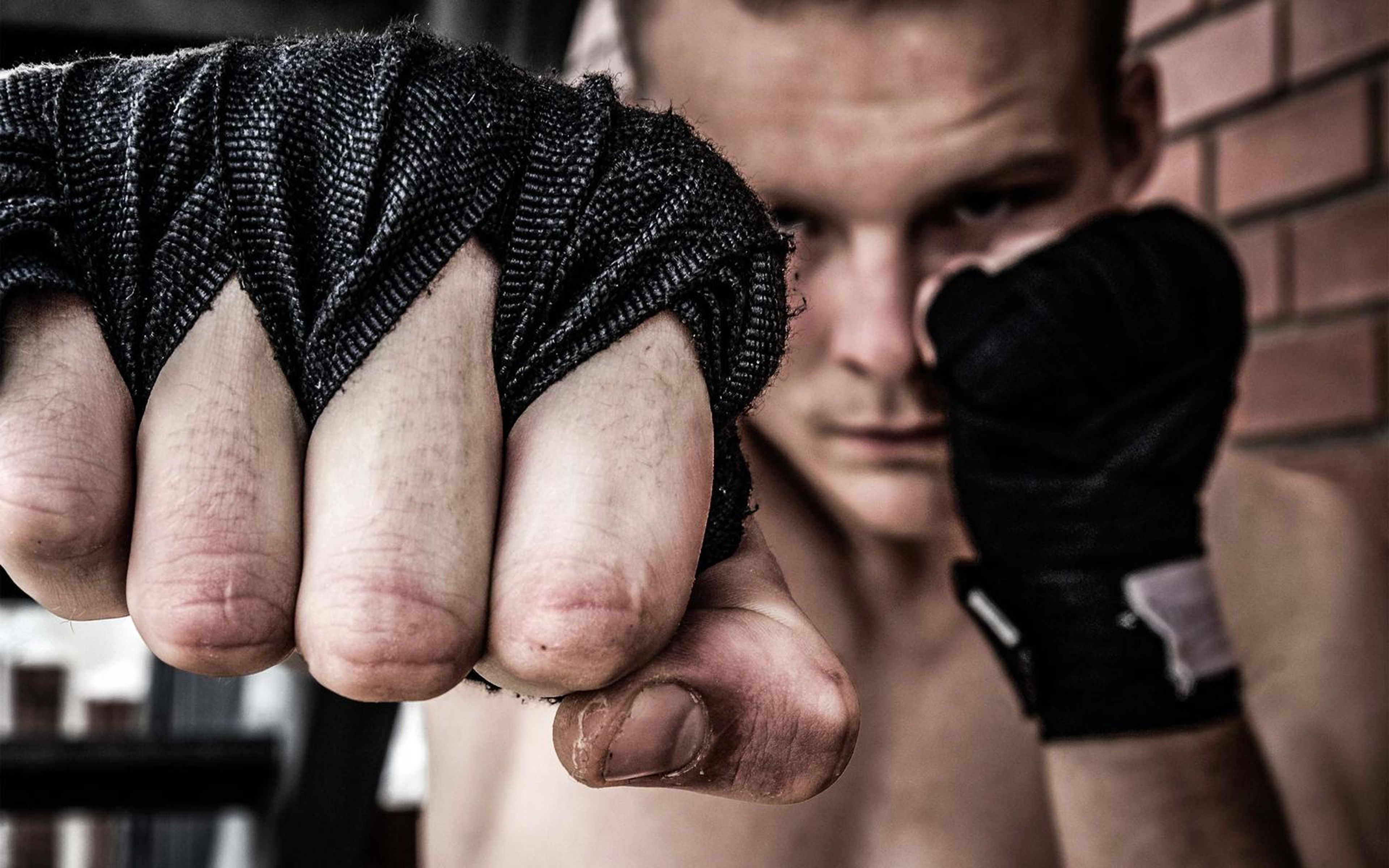 Fist in black hand wraps in sharp focus in front of the fighter's face, brick wall behind
