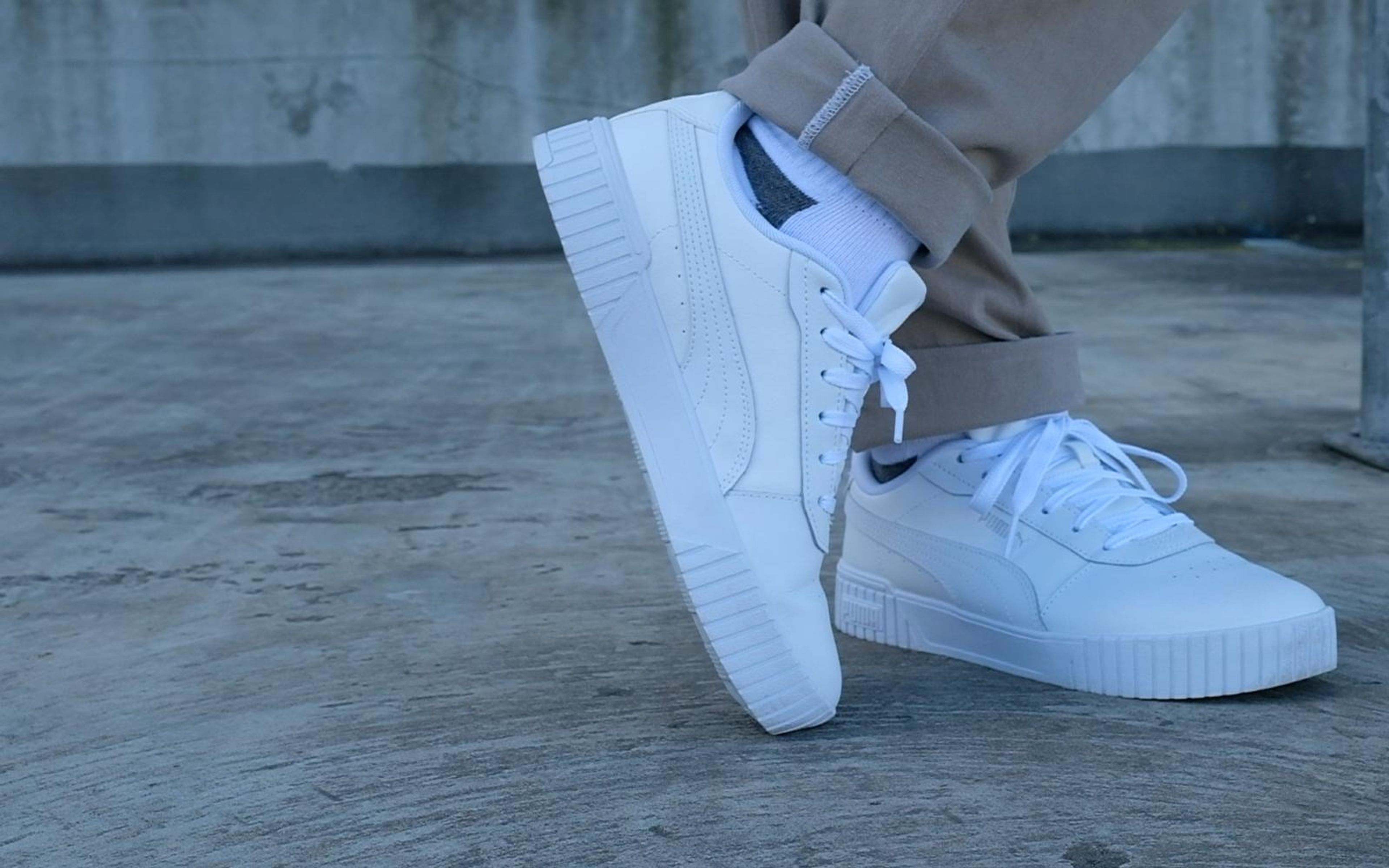 White leather sneakers on a concrete rooftop, one foot tilted on its side, tan cuffed trousers