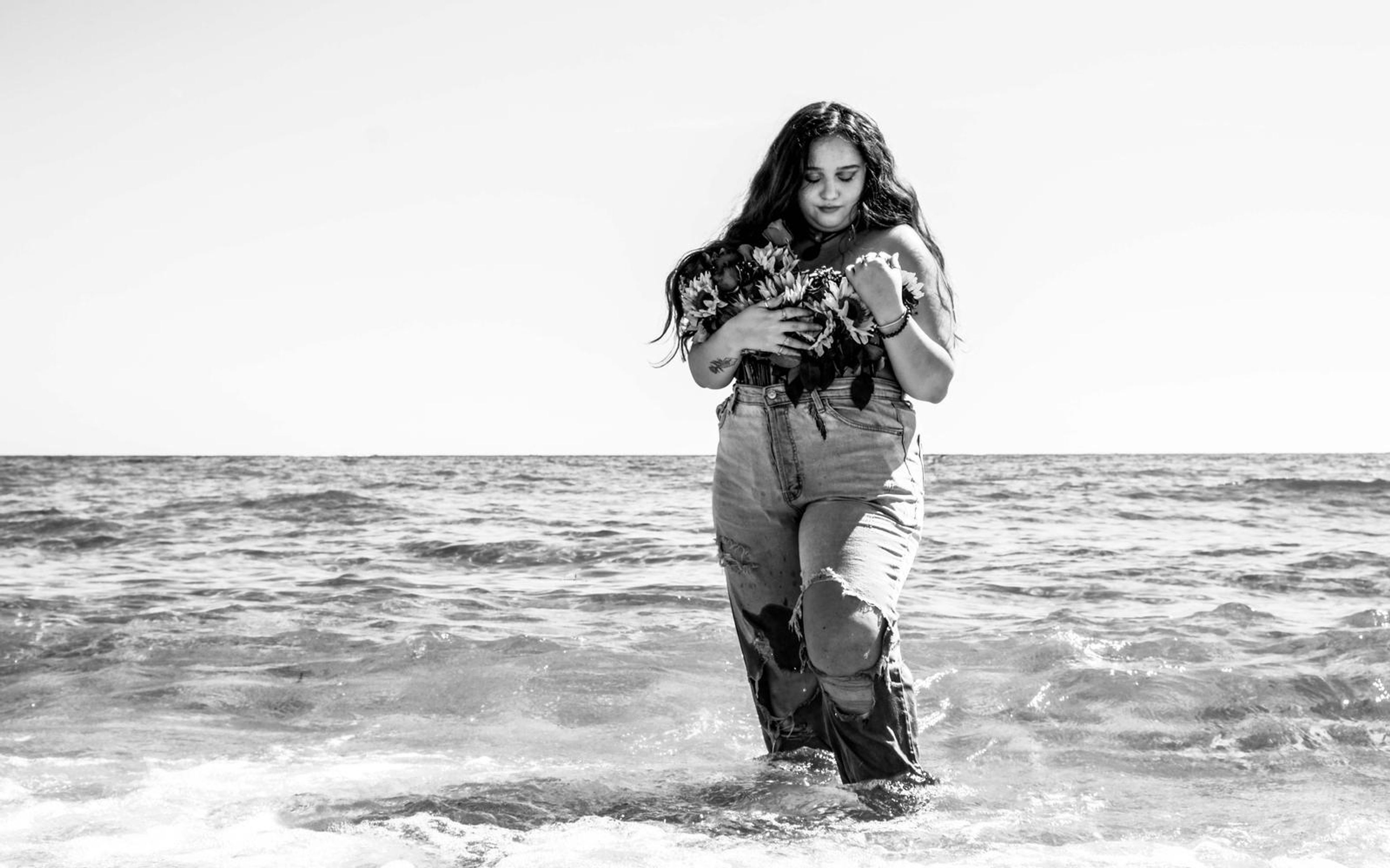 Black and white image of model wading in shallow sea, bouquet at chest, bright washed-out sky