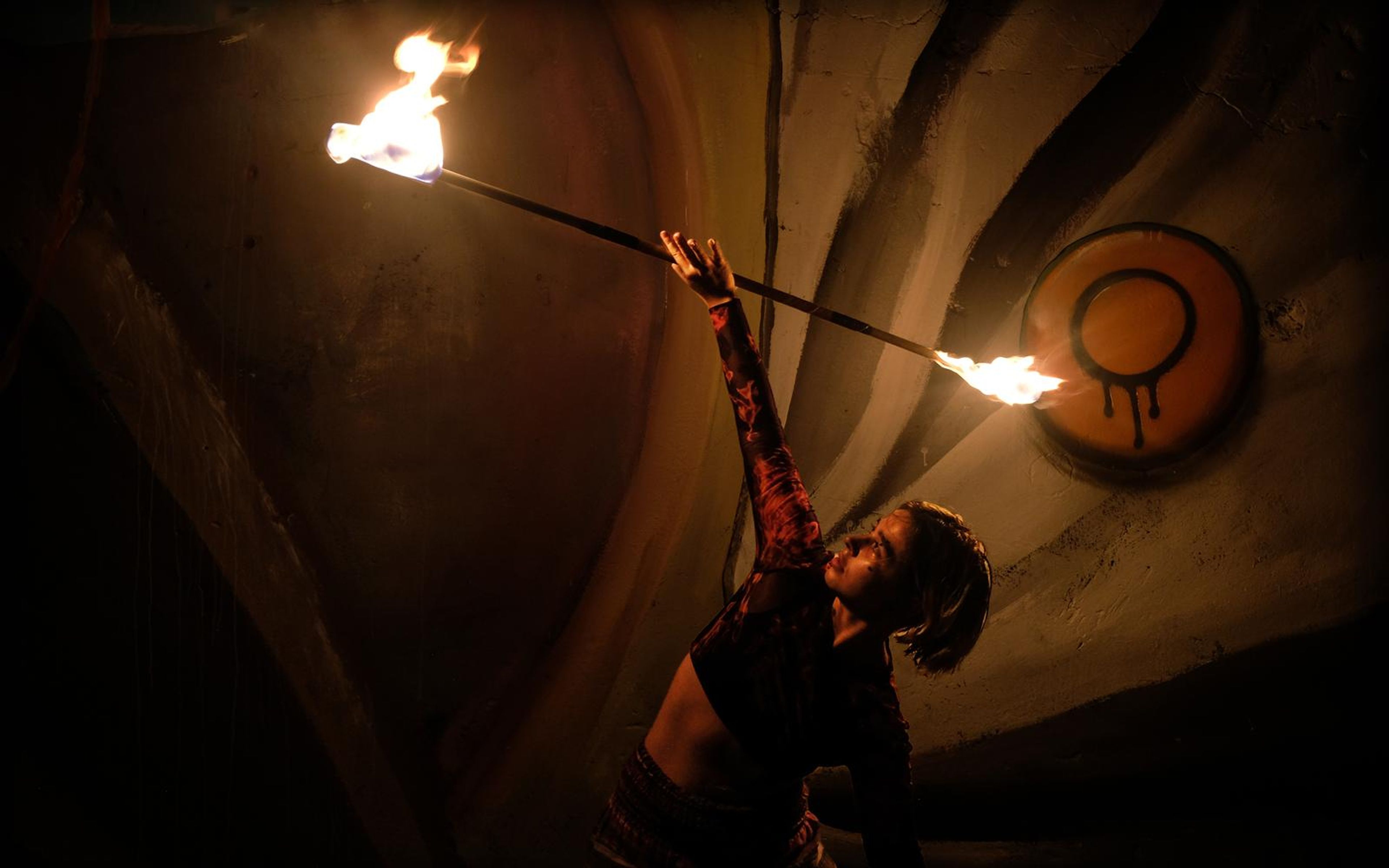Performer reaching up with a long fire staff, flames at both ends, orange emblem on the wall behind