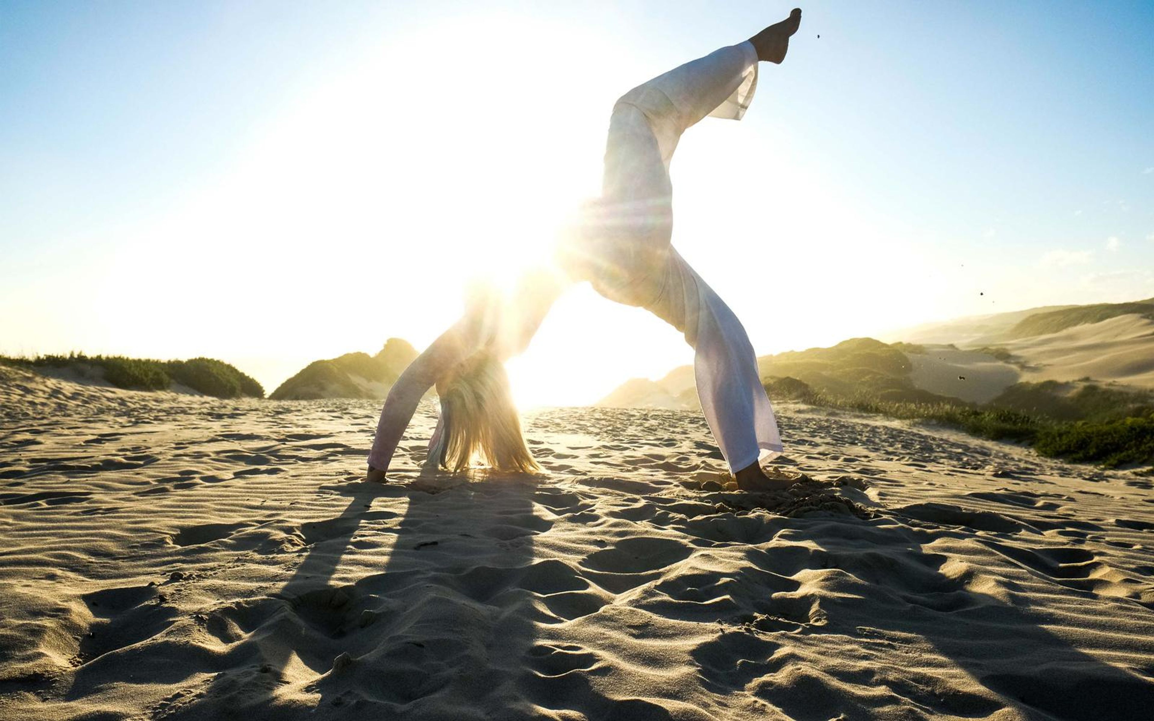 Silhouette of a woman in a handstand-style pose on dune sand, sun flaring behind
