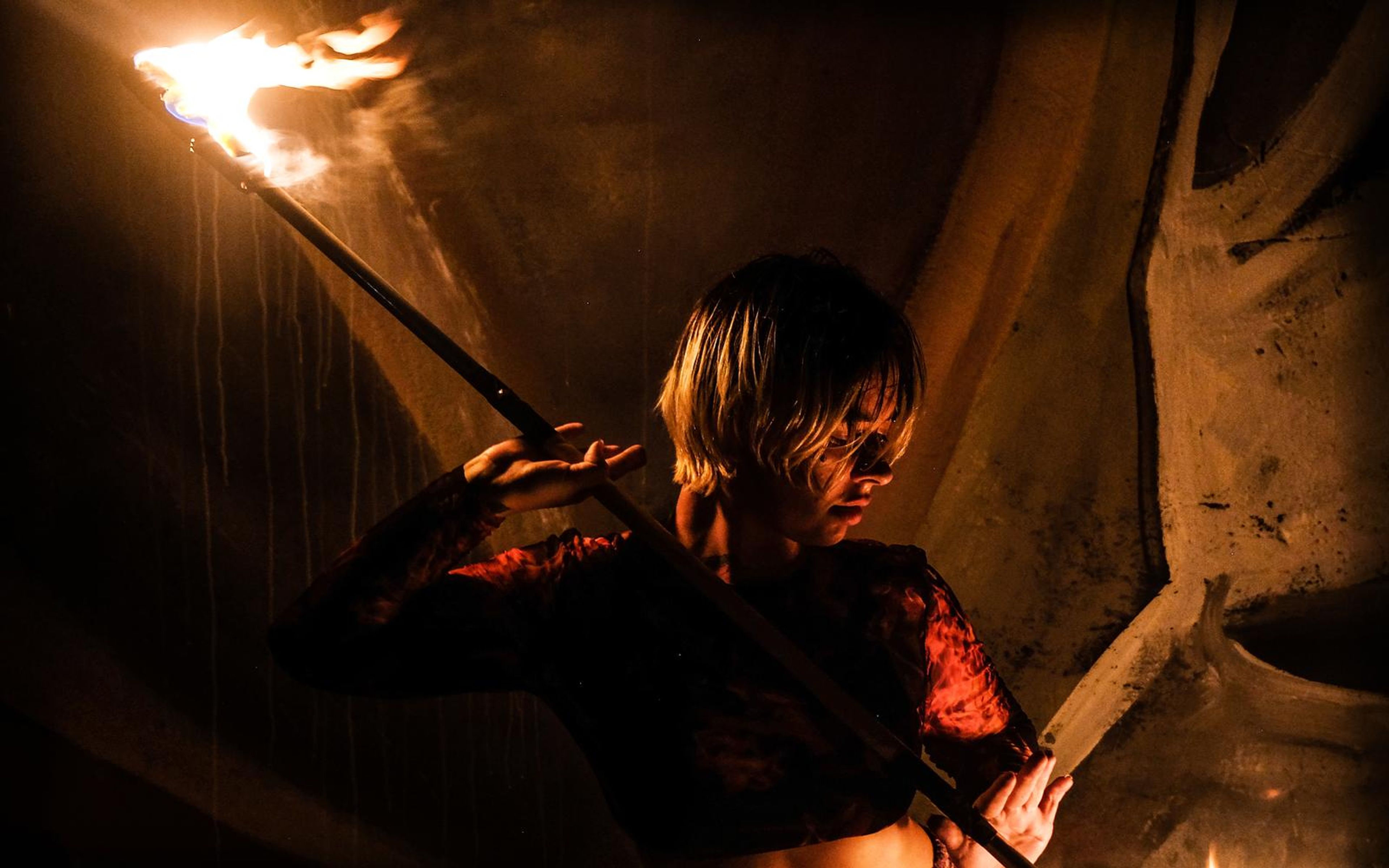 Performer in a red patterned top angling a long fire staff over one shoulder, flame at the top left