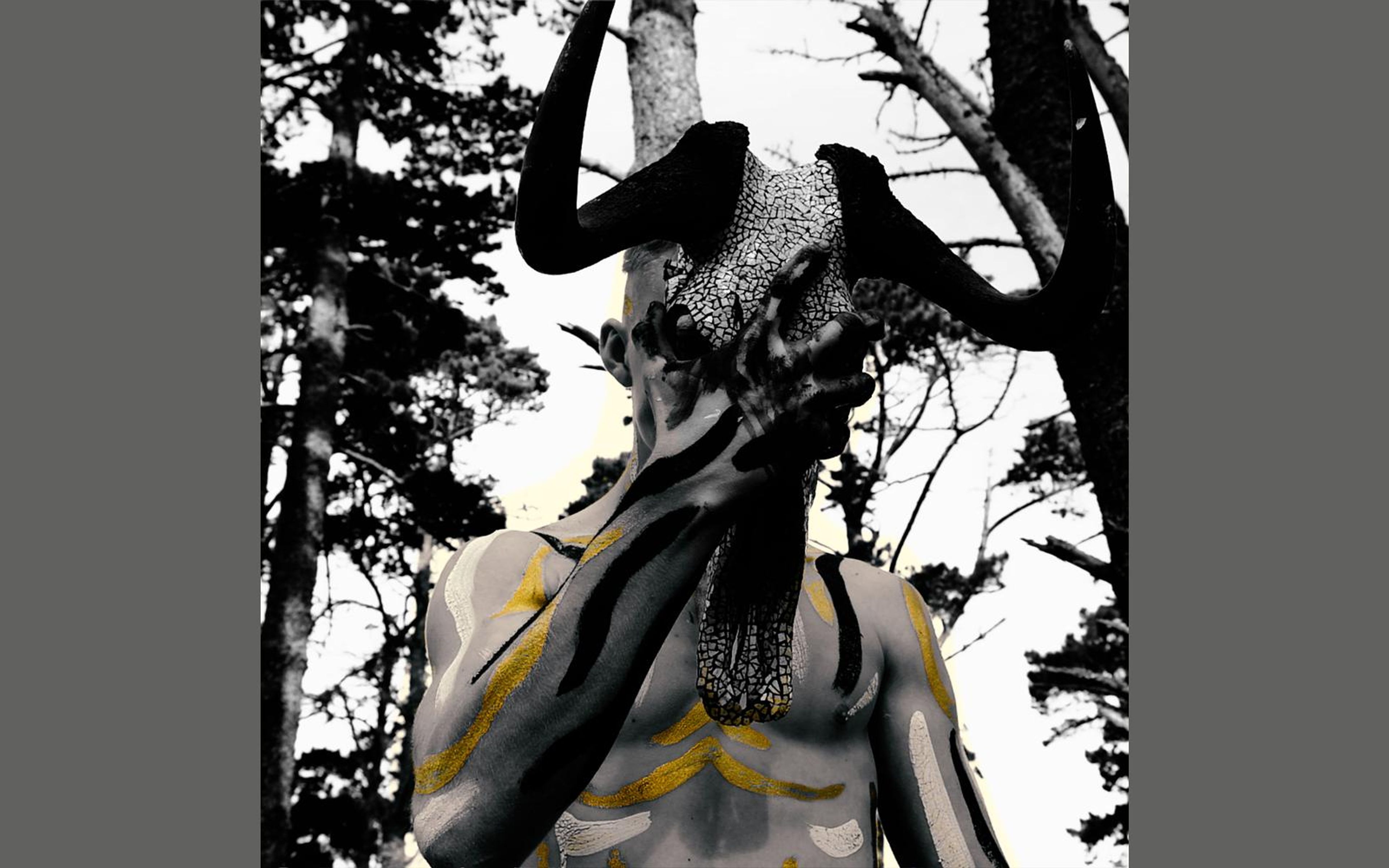 Performer with a horned skull mask and painted torso against pale sky and pine silhouettes