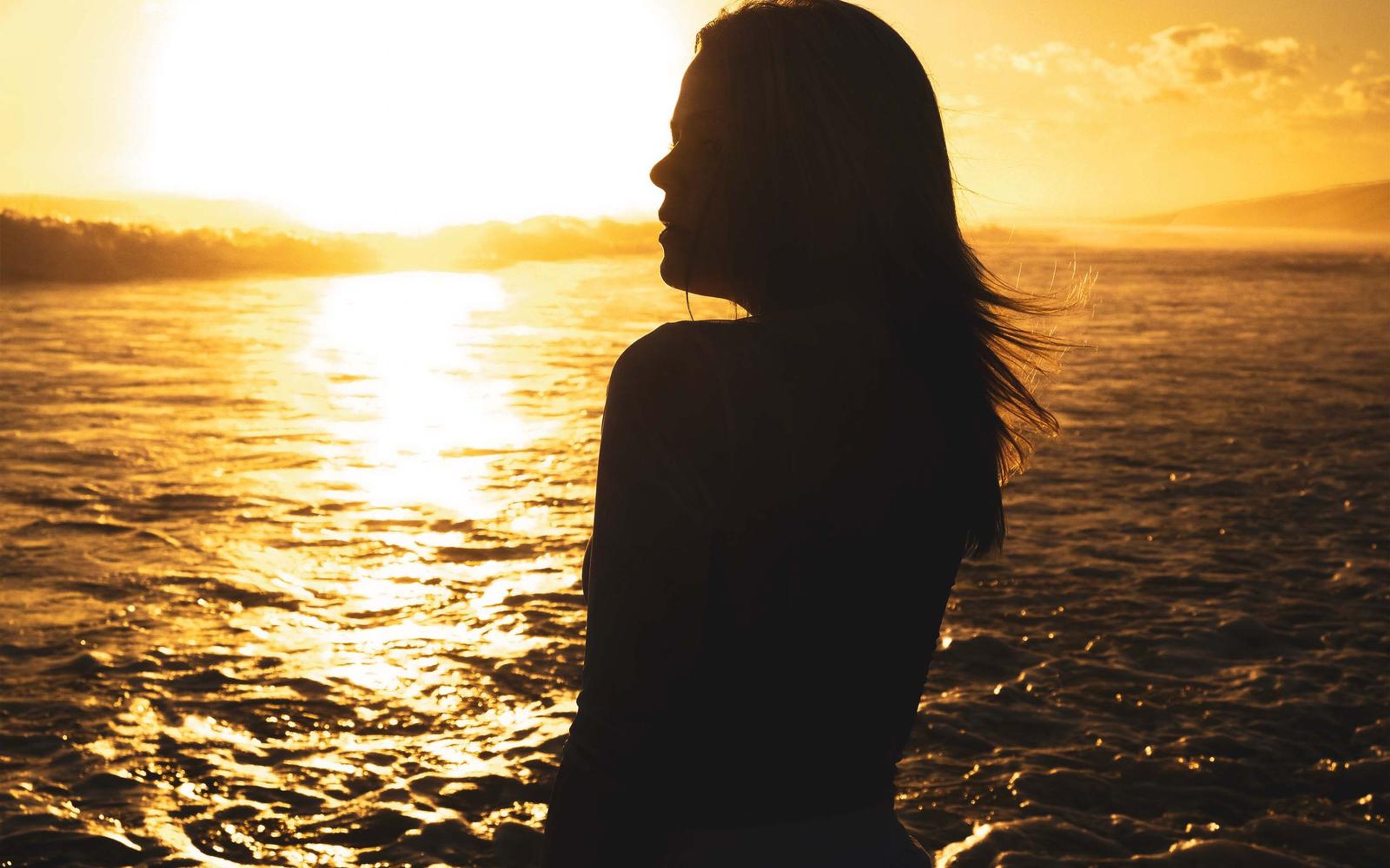 Woman in profile at sunset, hair blowing, sea glowing orange behind