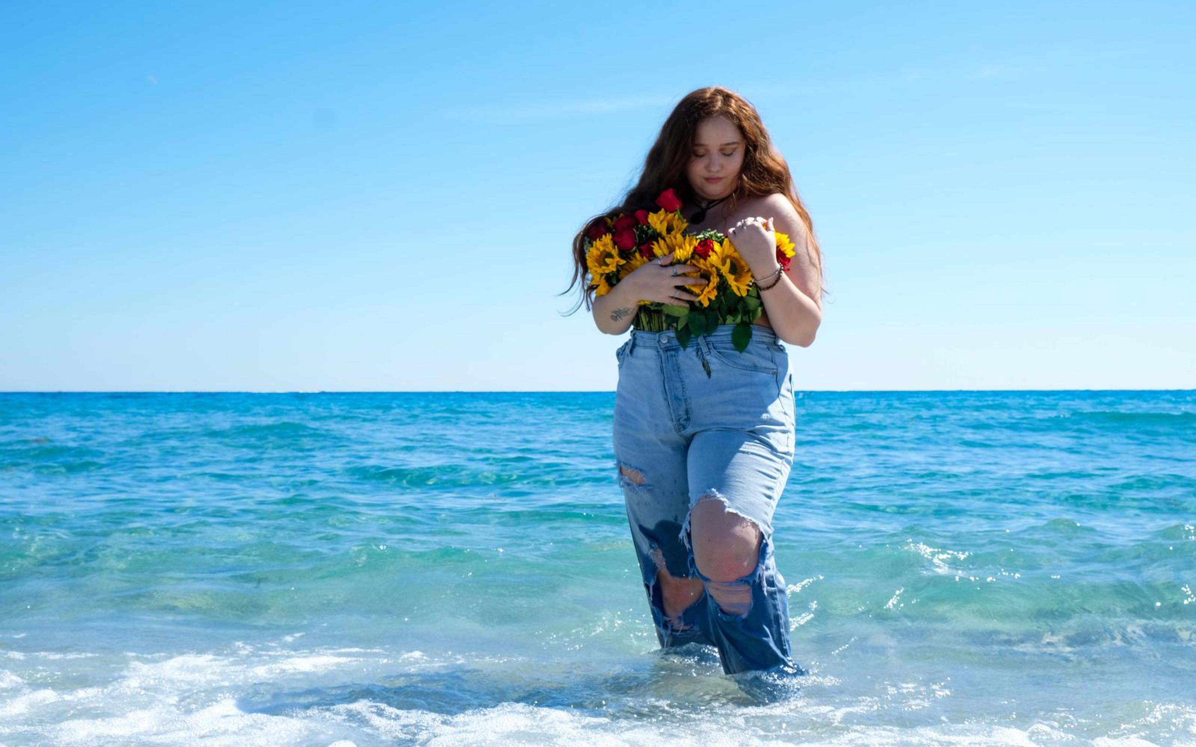 Model standing knee deep in blue sea holding the bouquet, torn denim legs, clear sky above
