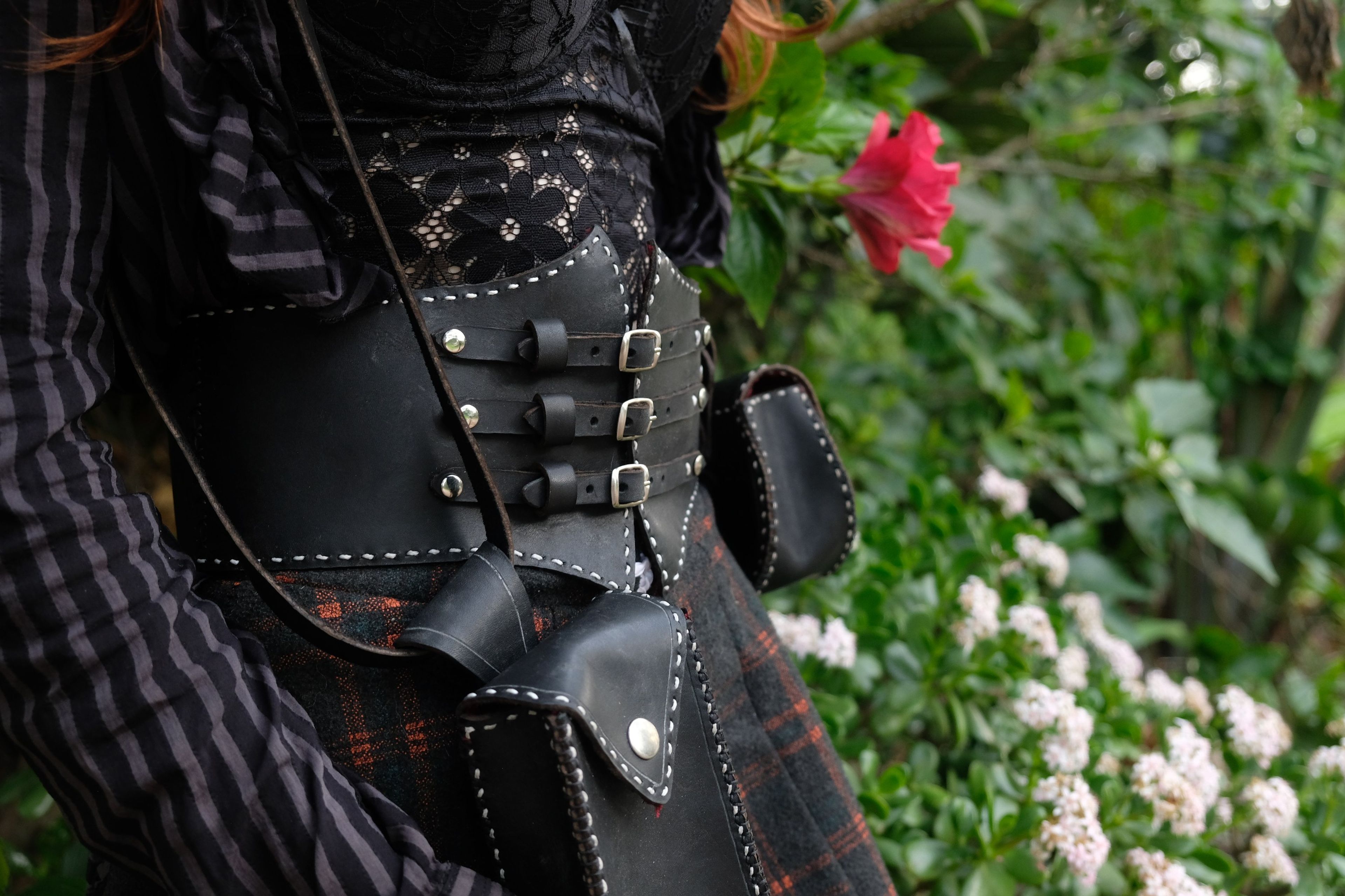 Black leather corset belt with buckles and a small shoulder bag beside a flowering bush