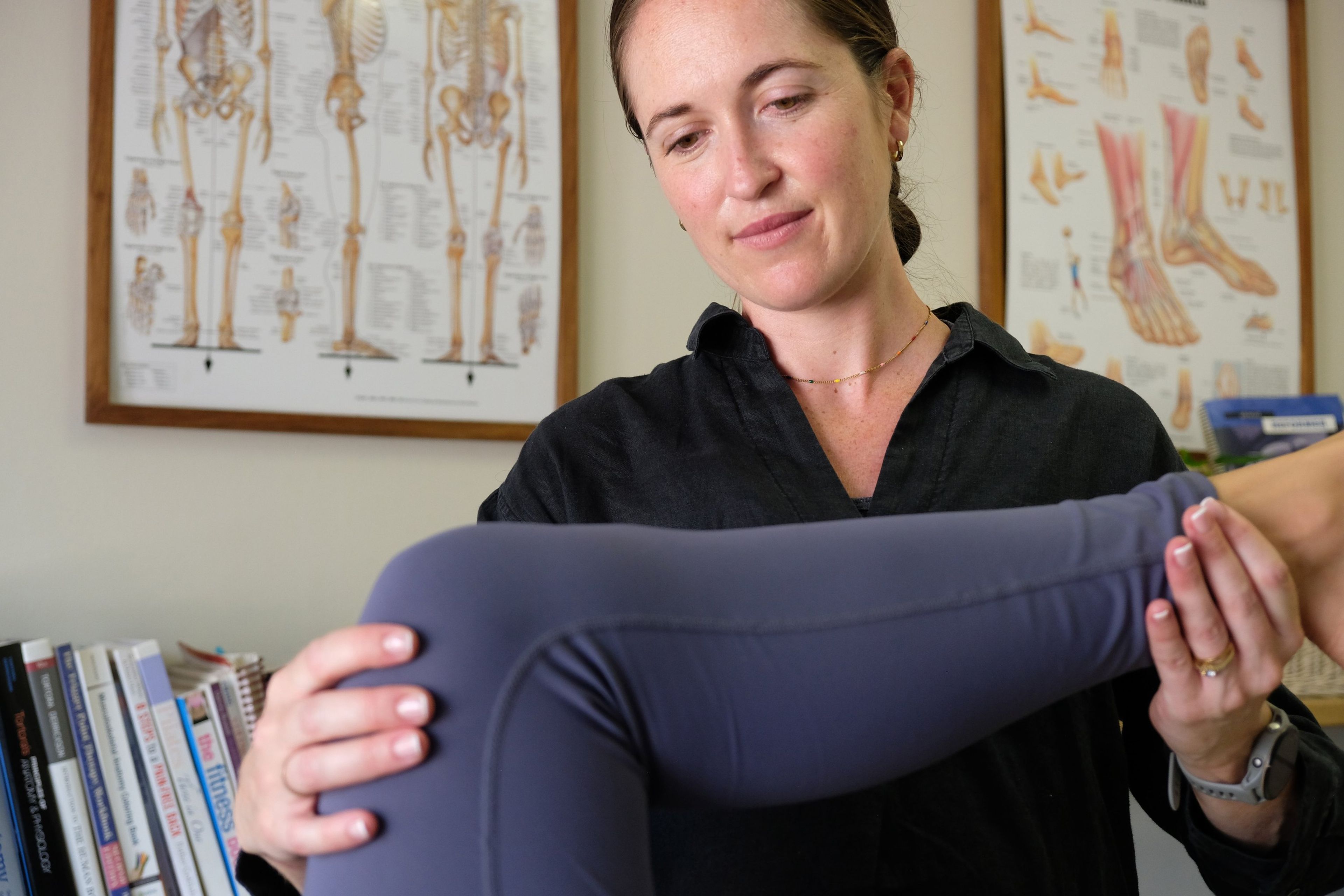 Team member guiding a client's raised leg during a treatment session, anatomy charts behind her