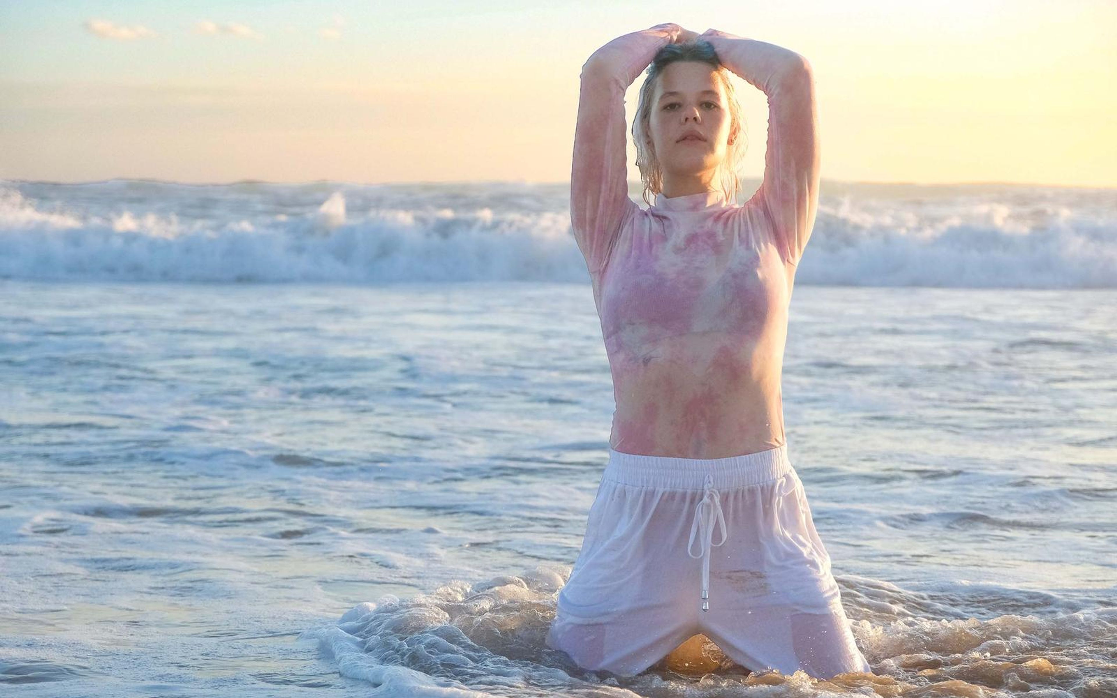 Woman kneeling in the waves with both arms raised over her head, wet pink top, soft pastel sky