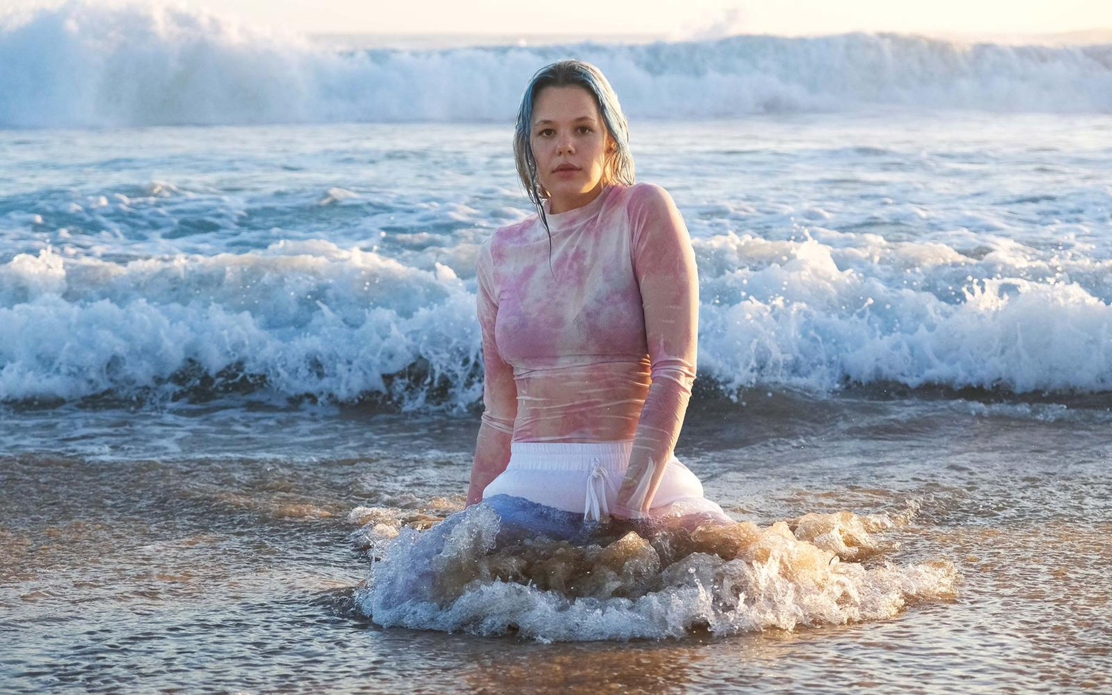 Woman kneeling in a breaking wave that splashes around her waist, large wave rolling behind