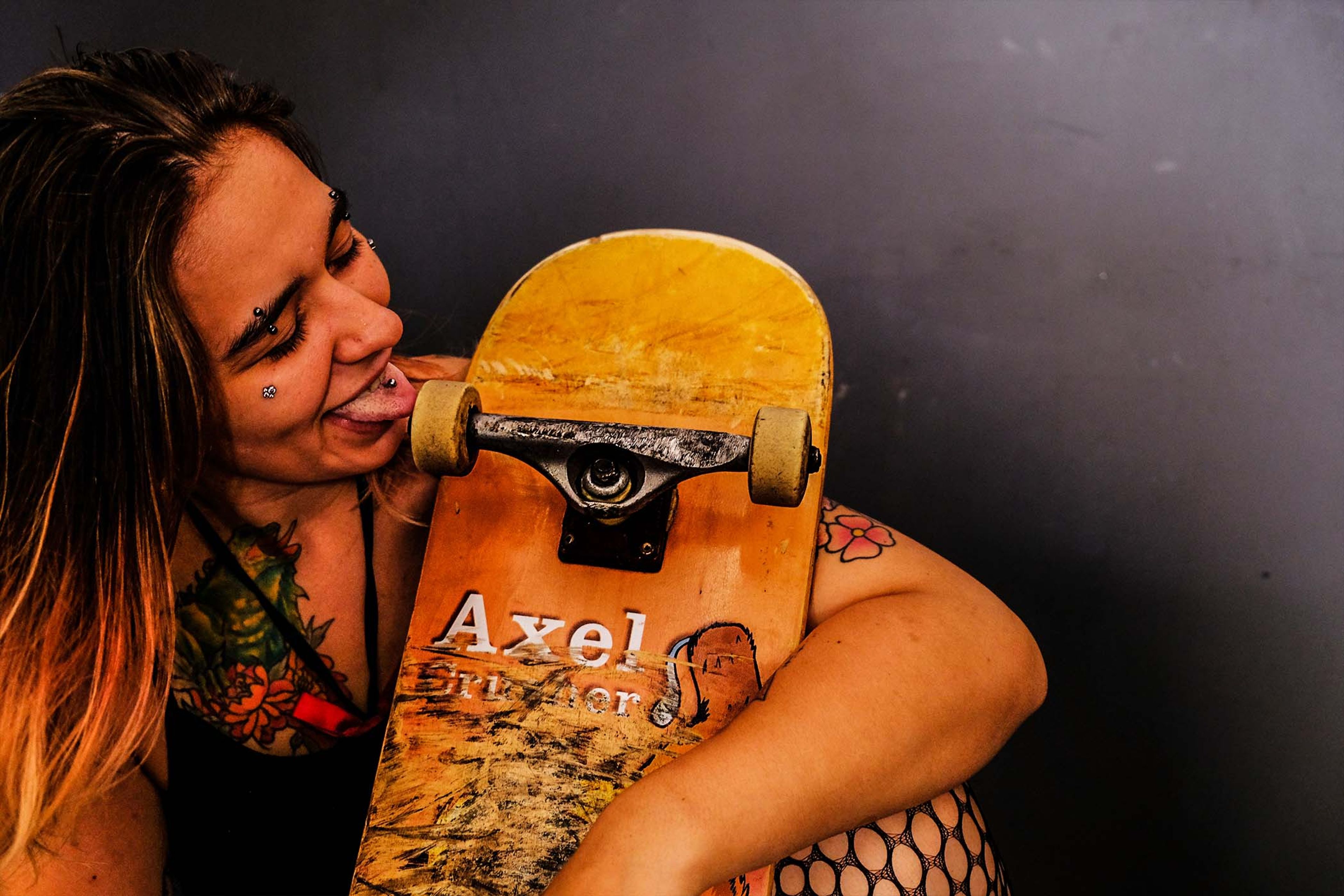 Subject holding a skateboard close to the face with tongue out, tattoos visible on the arm