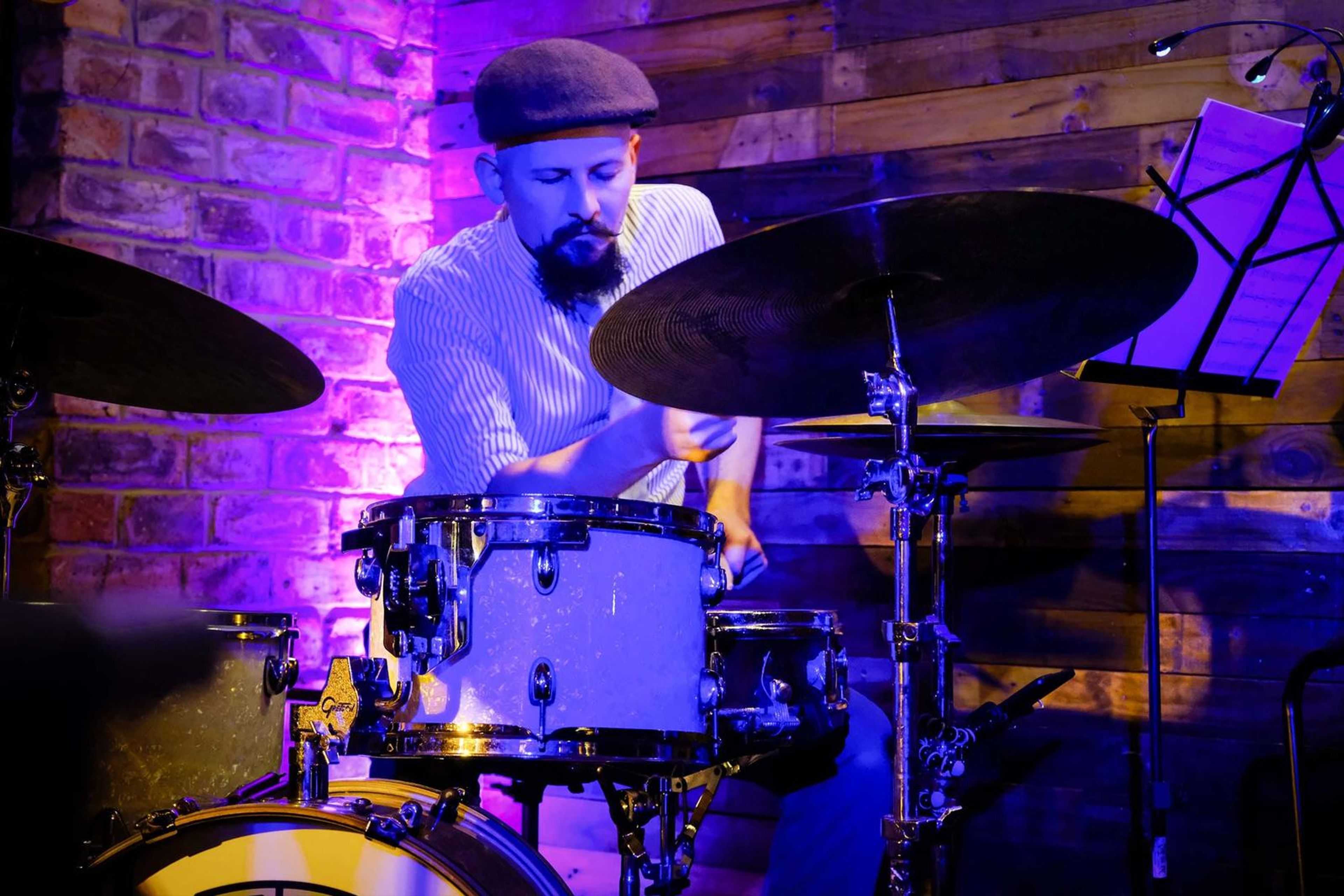 Drummer in a flat cap playing behind a white kit under purple light