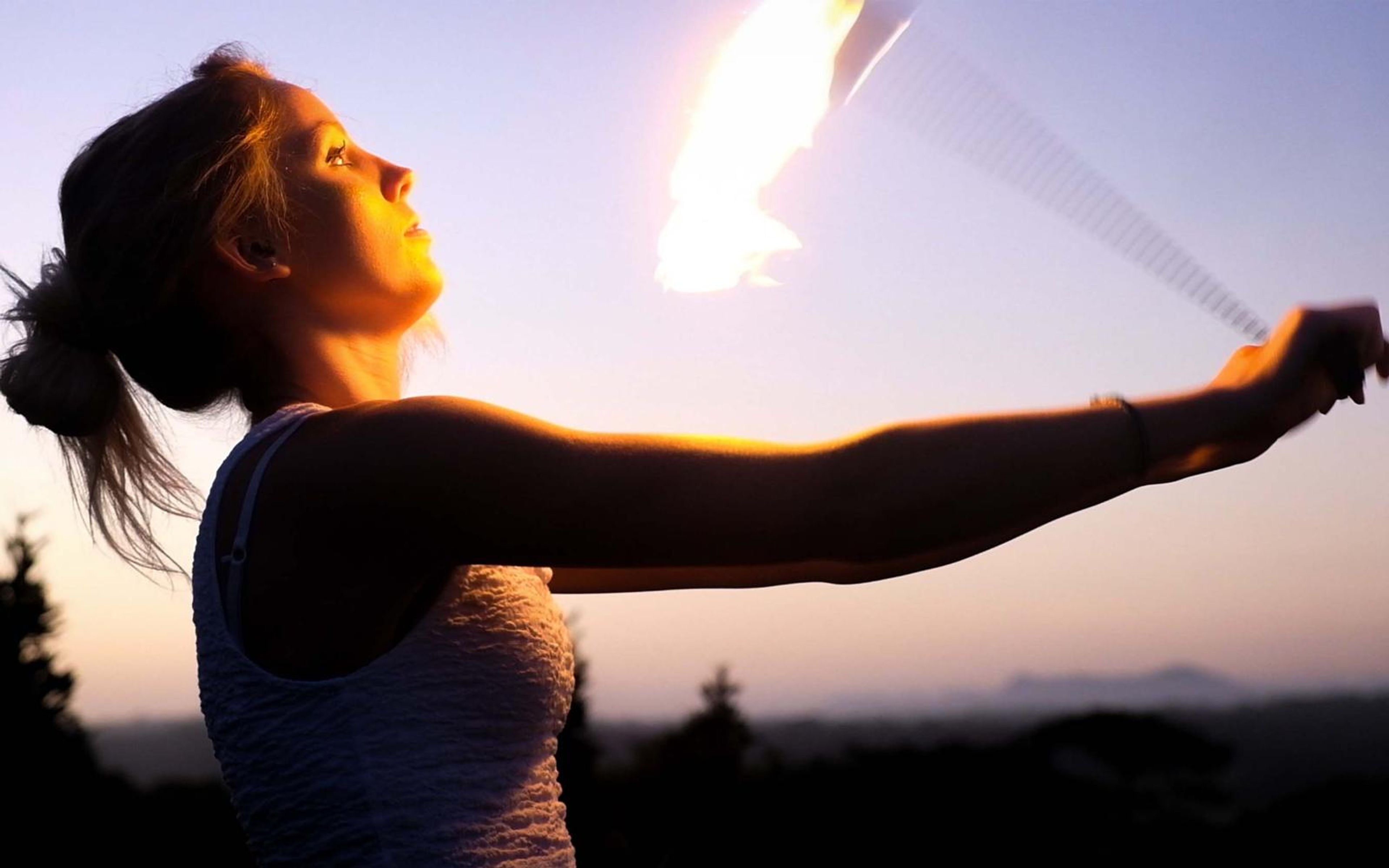 Woman with a ponytail swinging a flaming chain with arms outstretched, sunset glow behind her