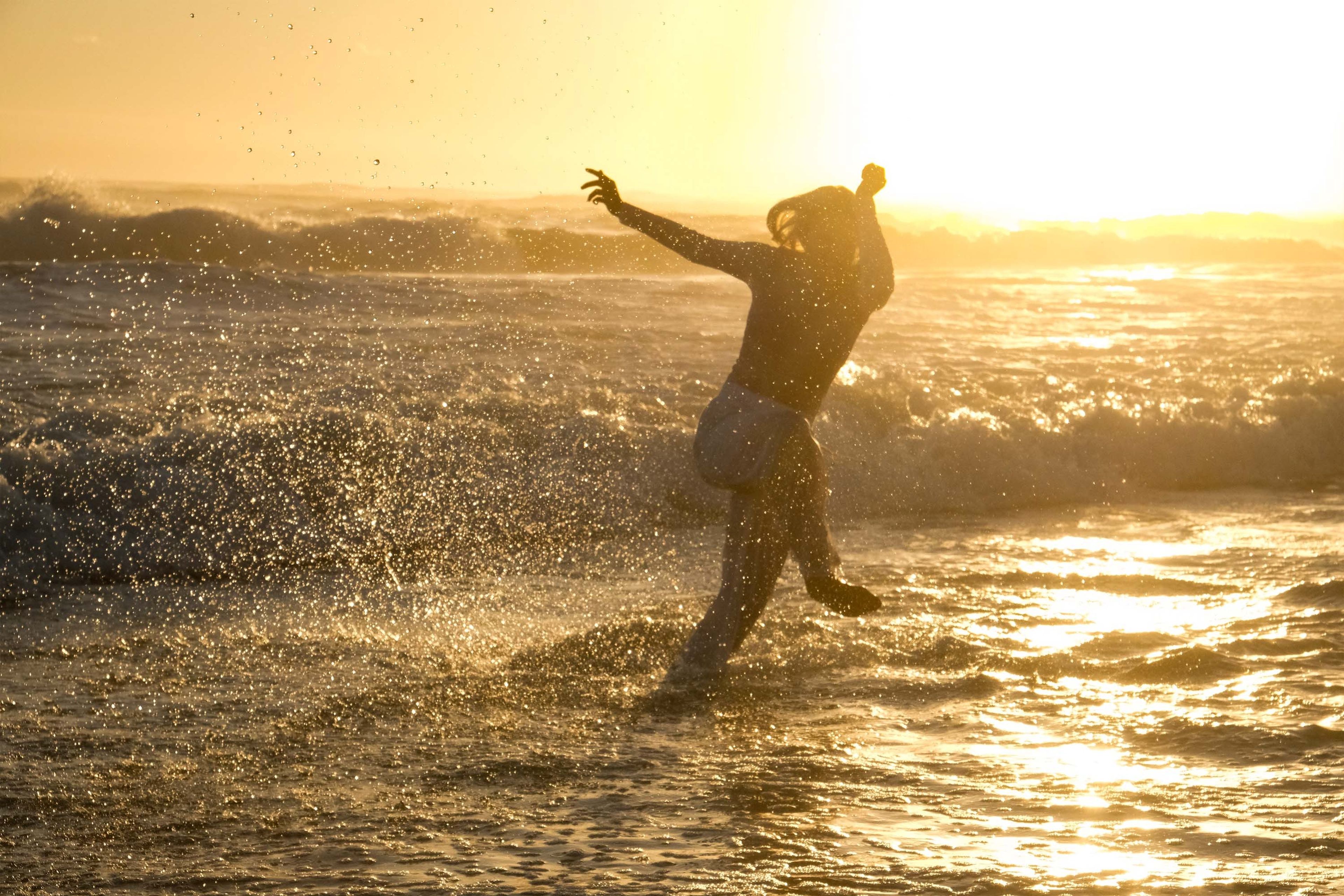 Silhouette of a woman walking into the sea toward a low setting sun, wet sand reflecting gold light