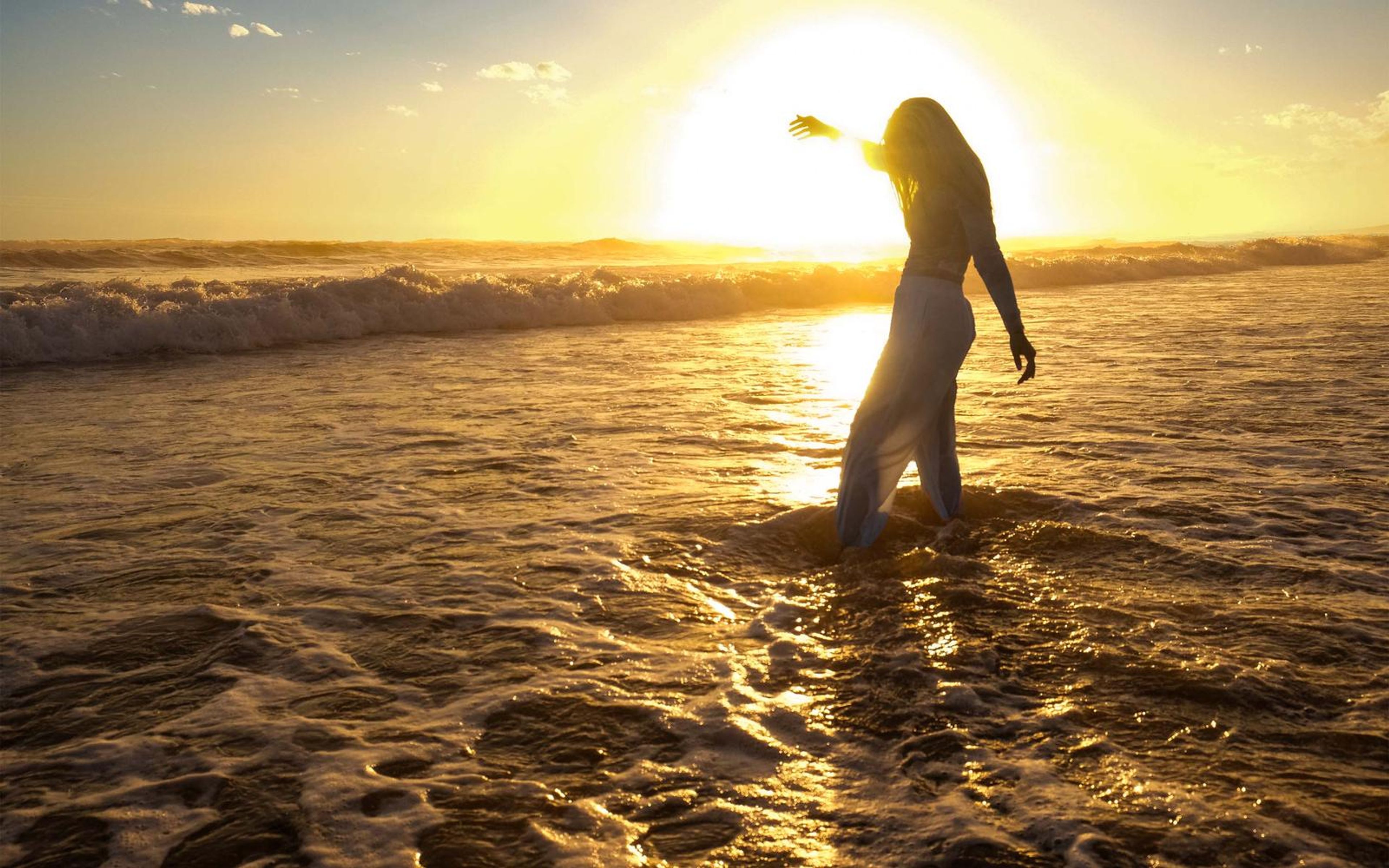 Woman in the shallows with one arm raised, backlit by a large setting sun
