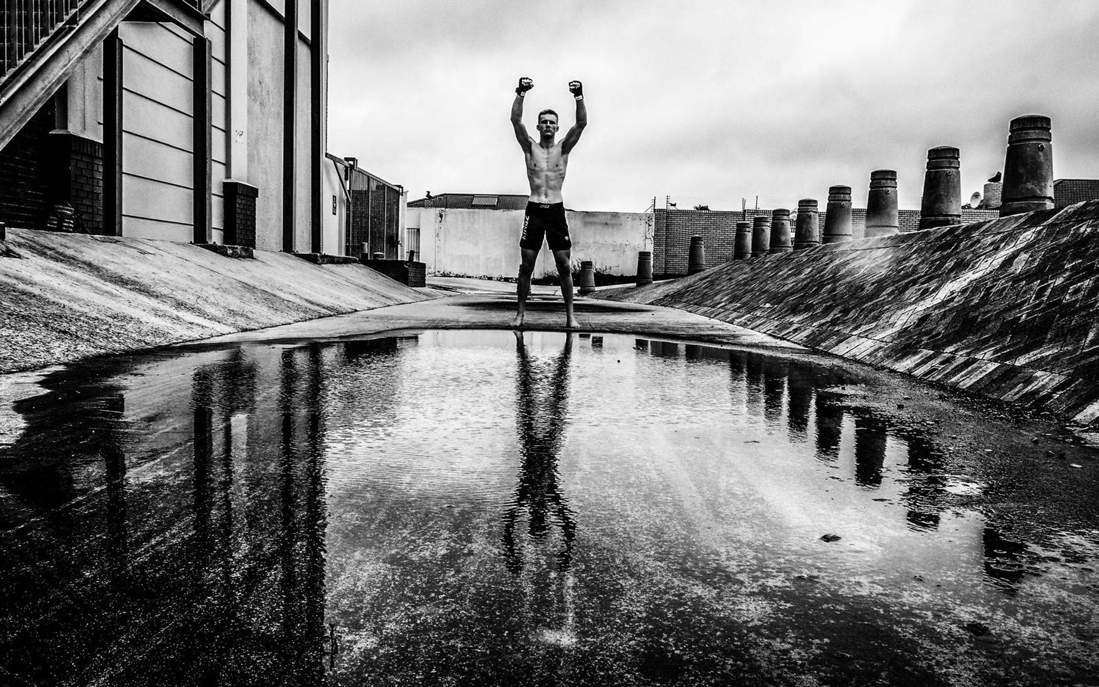 Black and white wide frame of a man with fists raised, reflected in a puddle, ramp and bollards behind