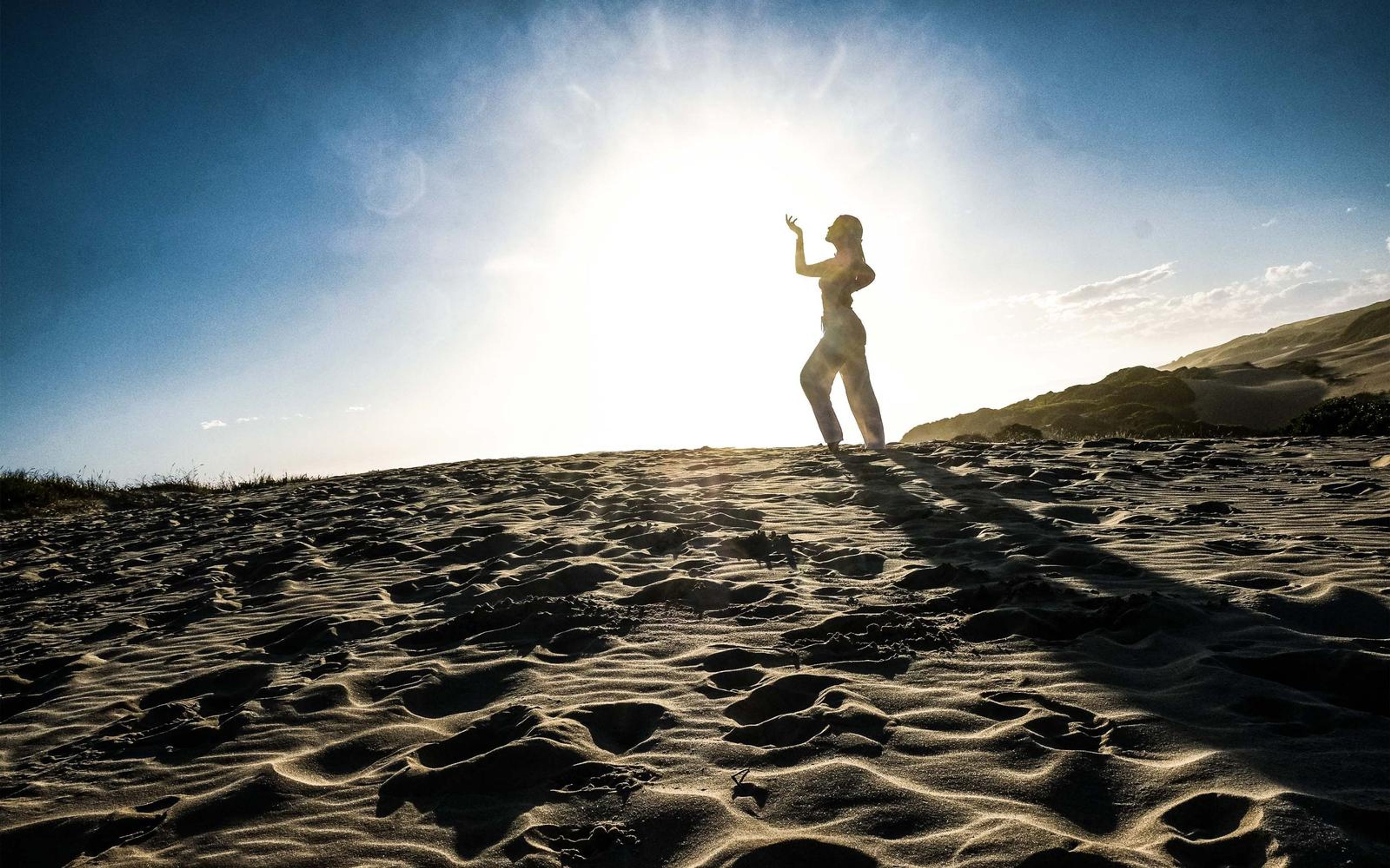 Small figure on a dune ridge raising her hand, sun burst behind in a blue and white sky