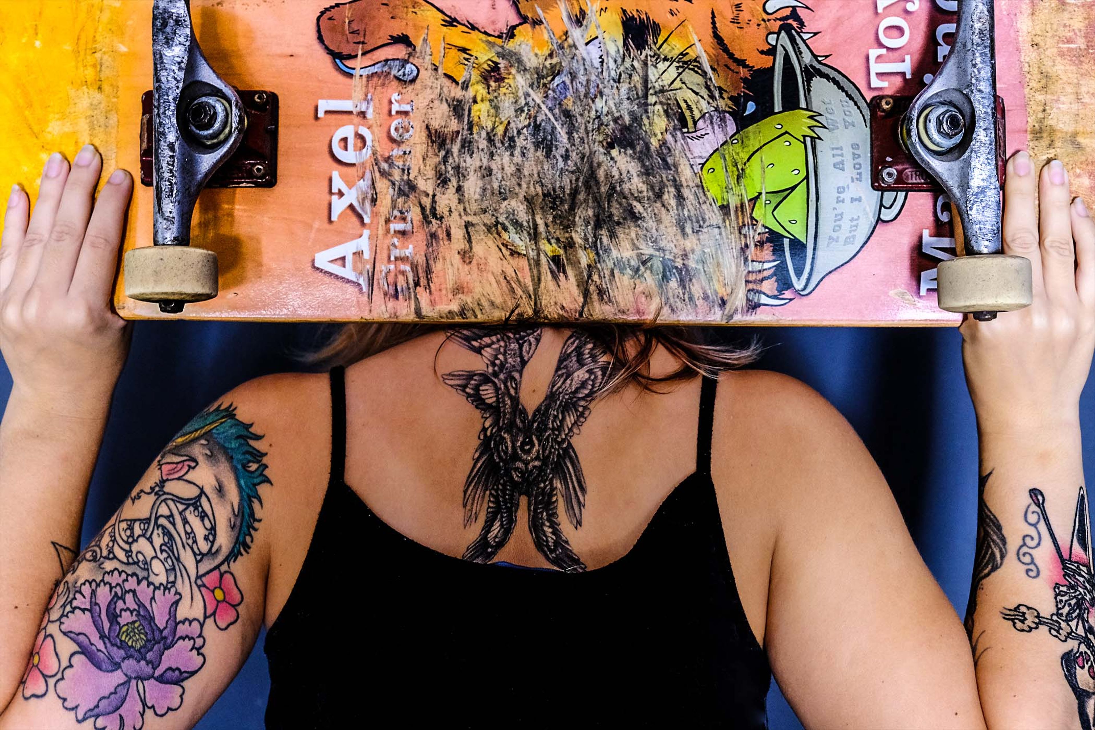 Overhead shot of a skateboard held above the head, back and arm tattoos visible