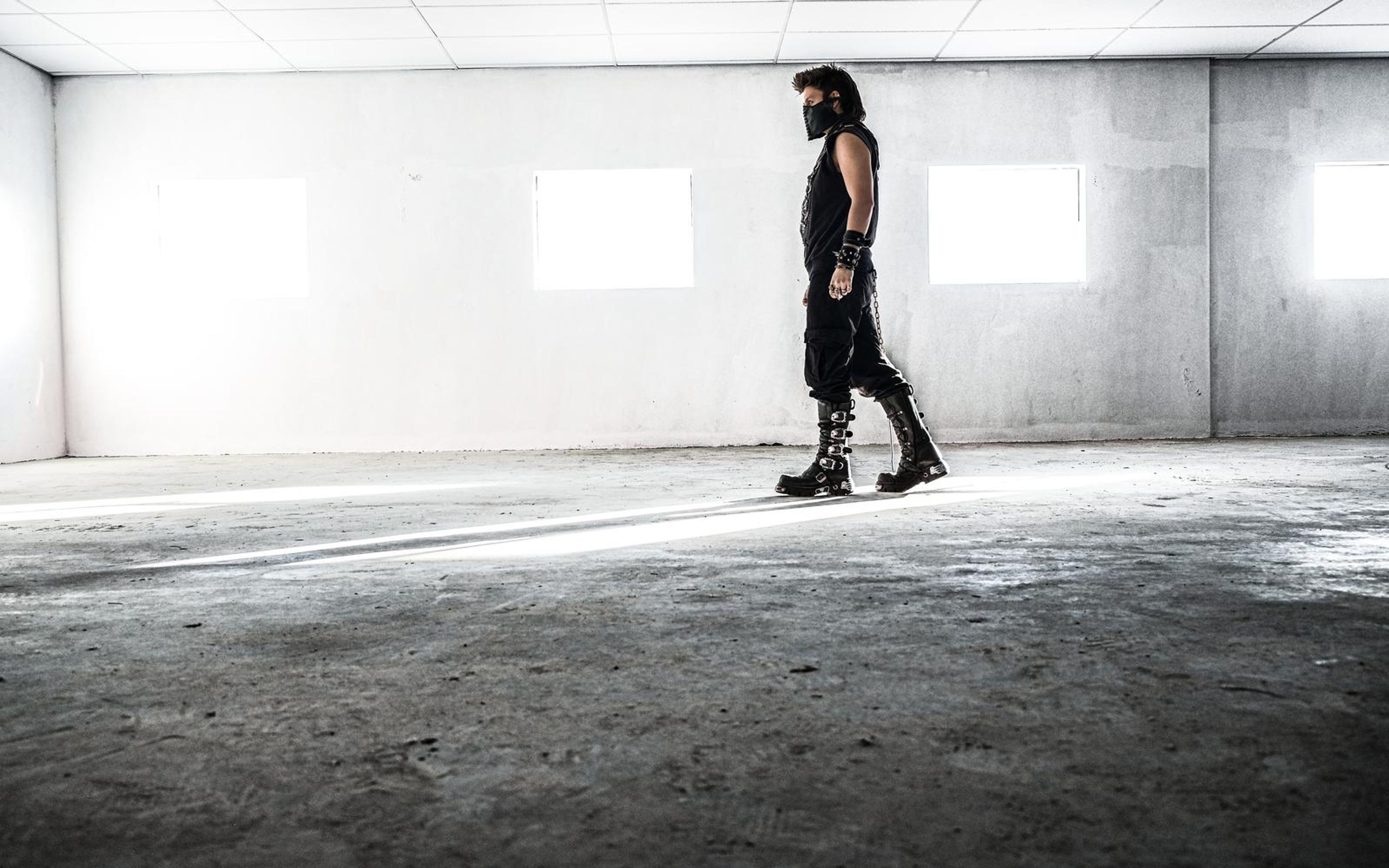 Masked man in black walks through a bare concrete warehouse, dark grade with bright windows and a light streak on the floor