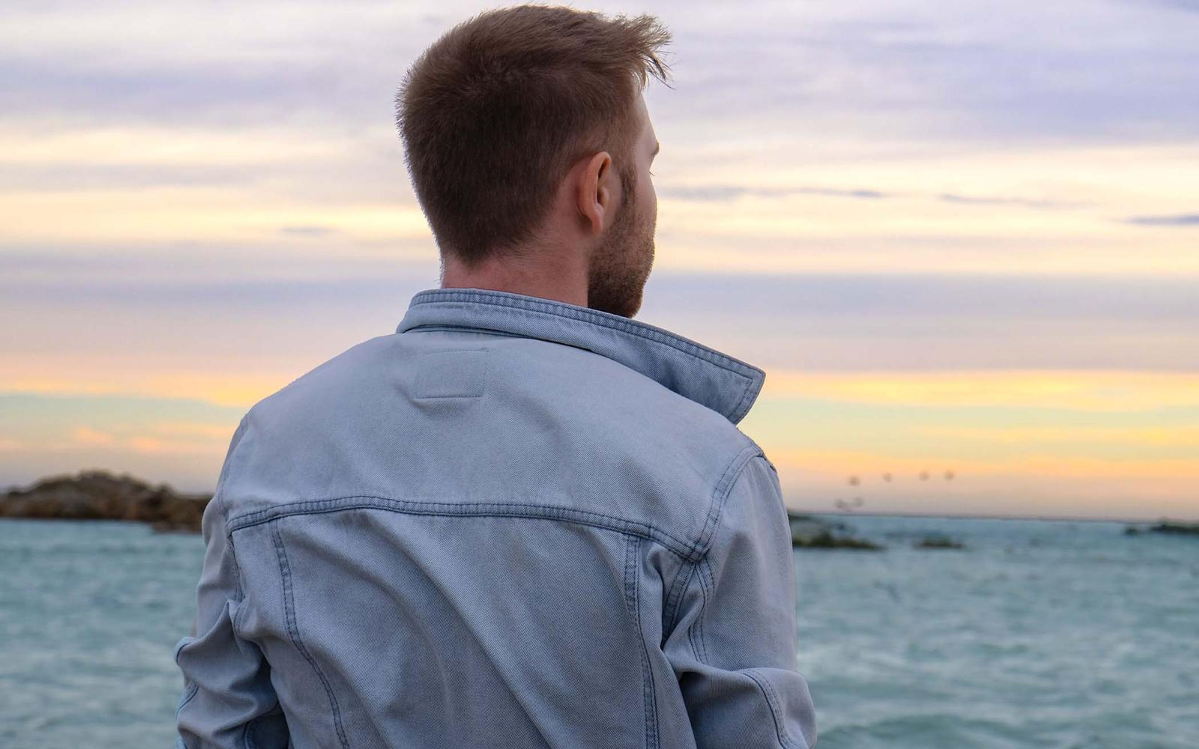 Man seen from behind in a denim jacket looking out over calm sea at dusk, pink and yellow sky