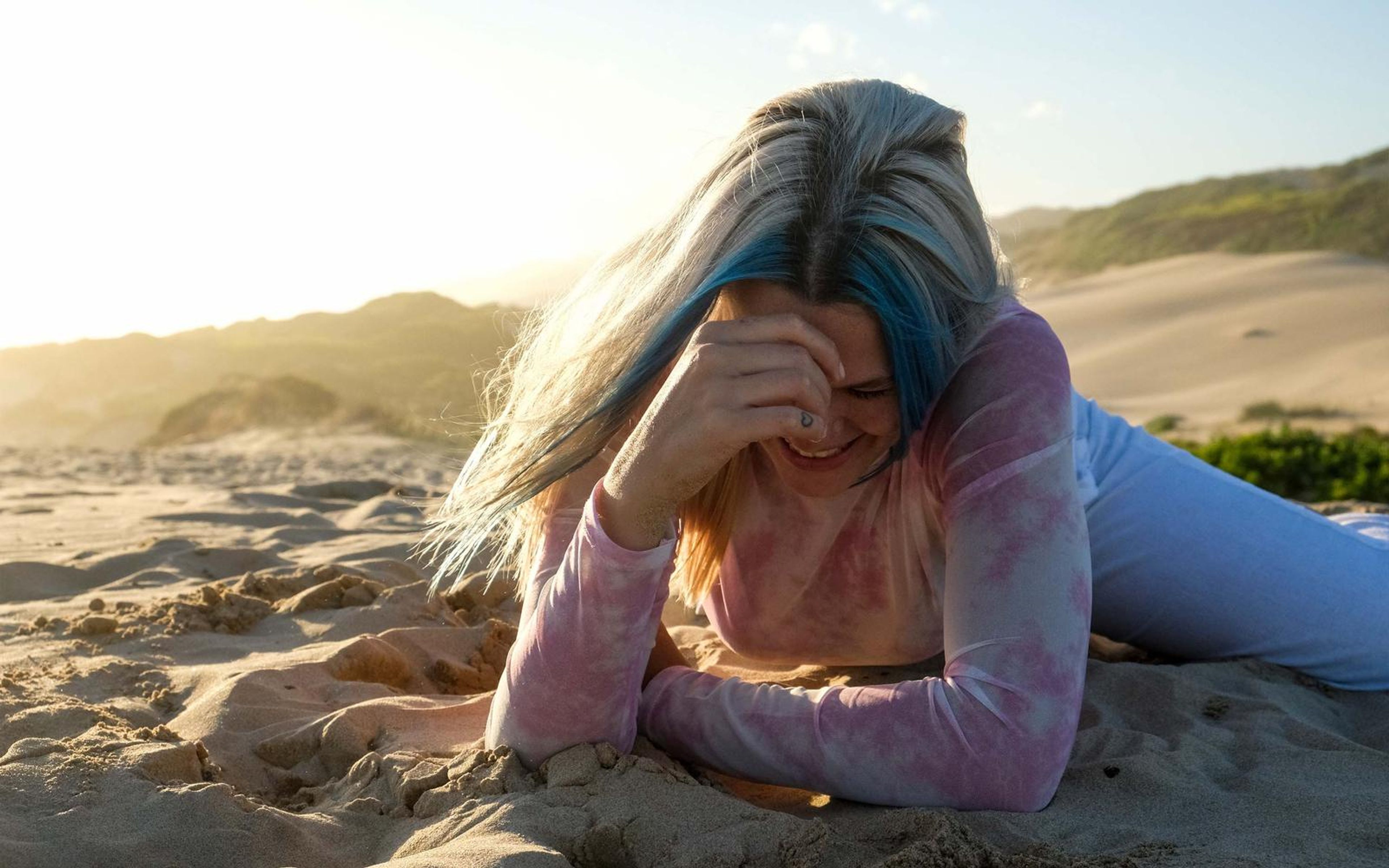 Woman lying on the sand laughing with a hand across her forehead and hair swept by wind
