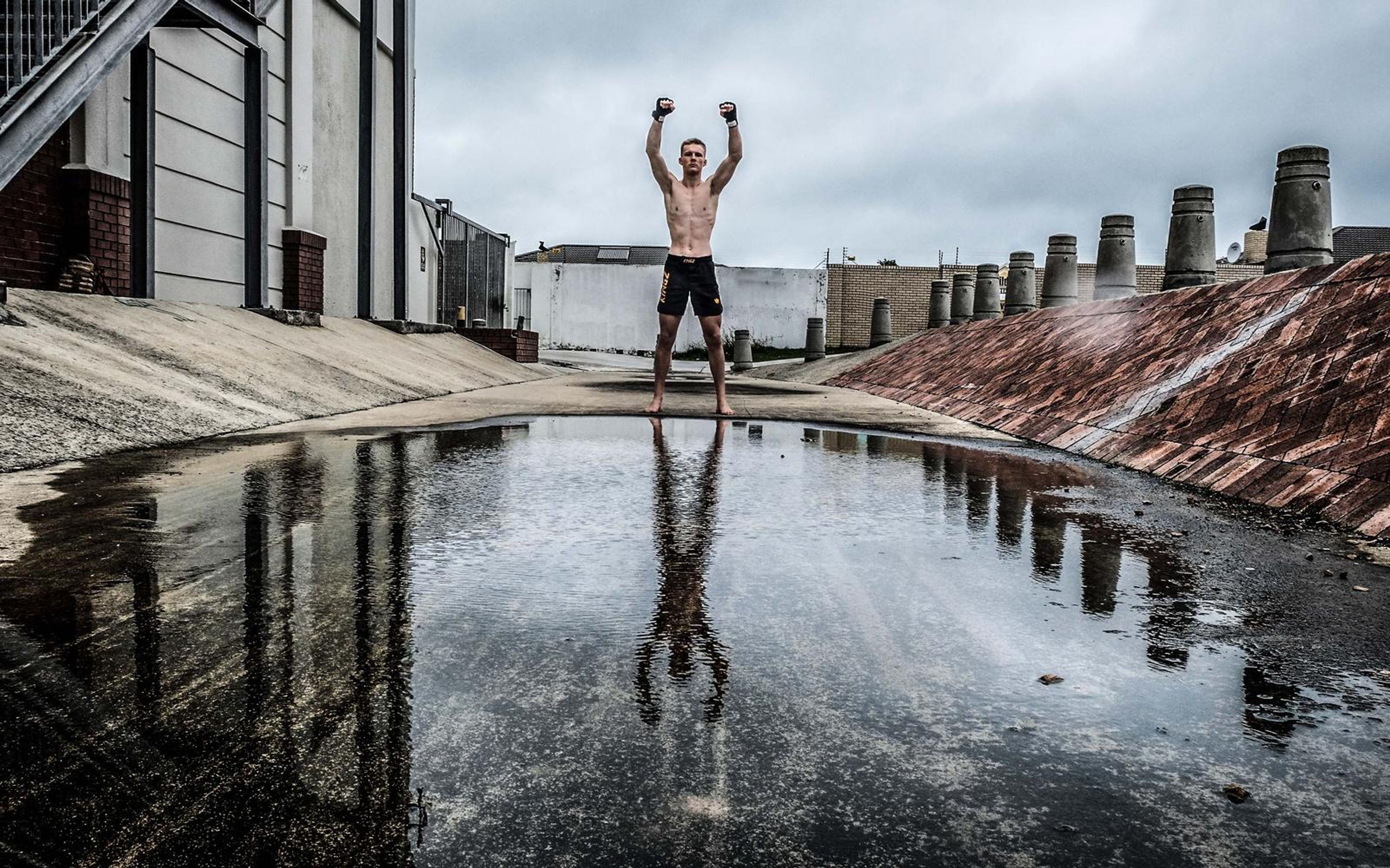 Man standing with arms raised beside a rain puddle that mirrors him, wet concrete and grey sky