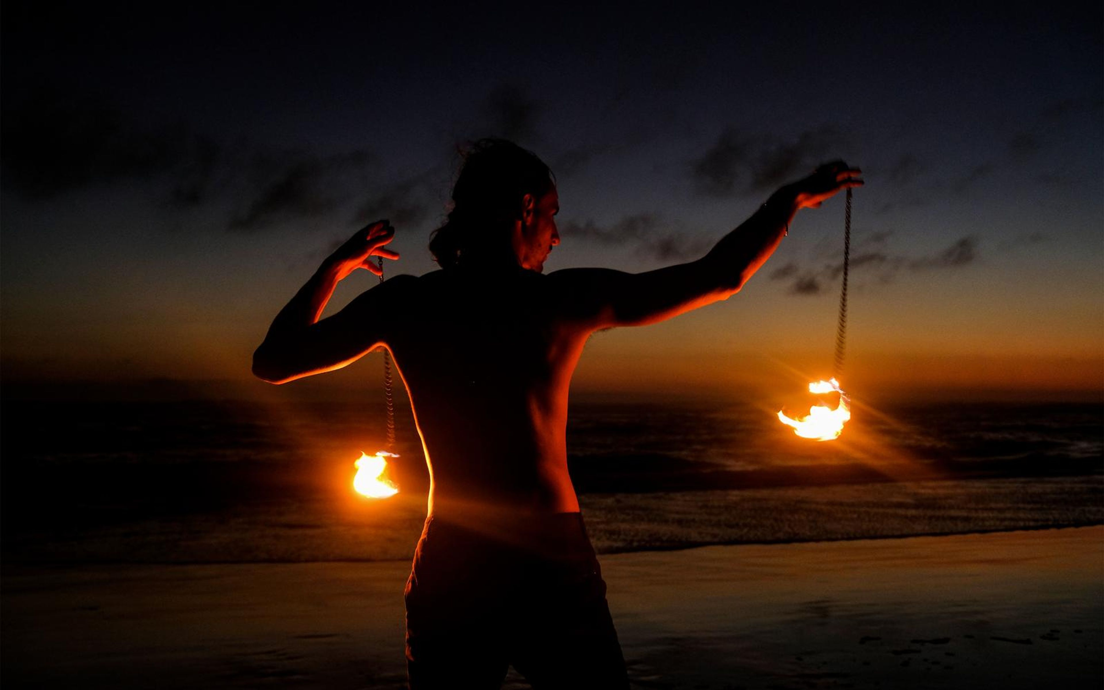 Performer silhouetted from behind on a beach holding two fire poi, orange light on the horizon
