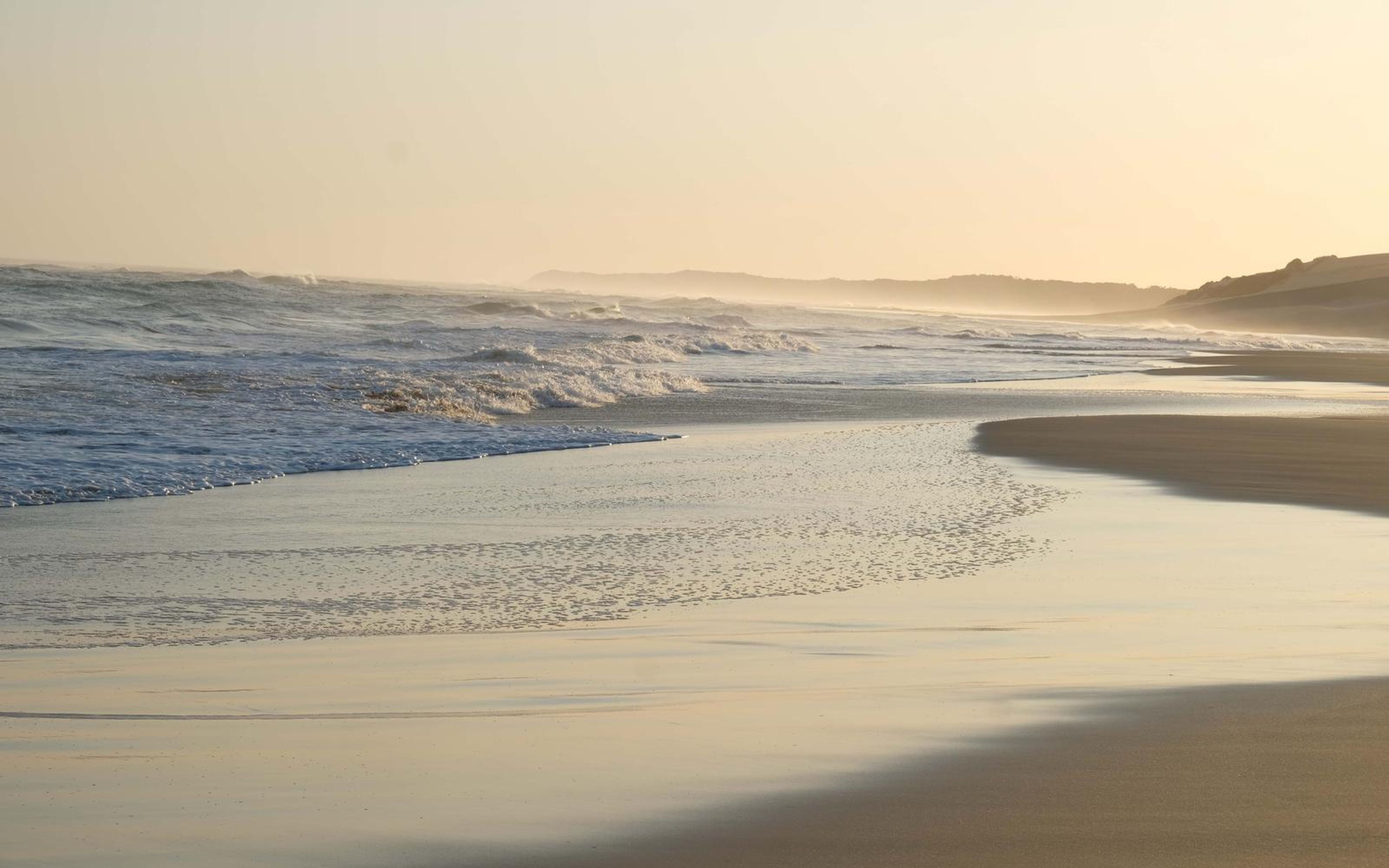 Curving shoreline with surf, a pale dune headland and warm haze at the far end