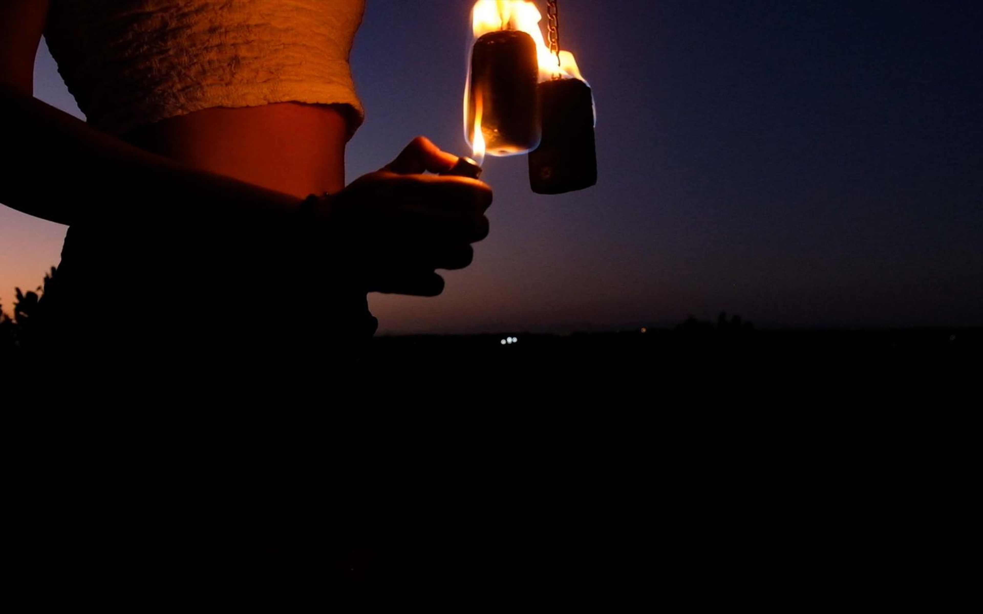 Wide crop of a hand lighting a fire prop on chains at dusk, with a dark horizon behind