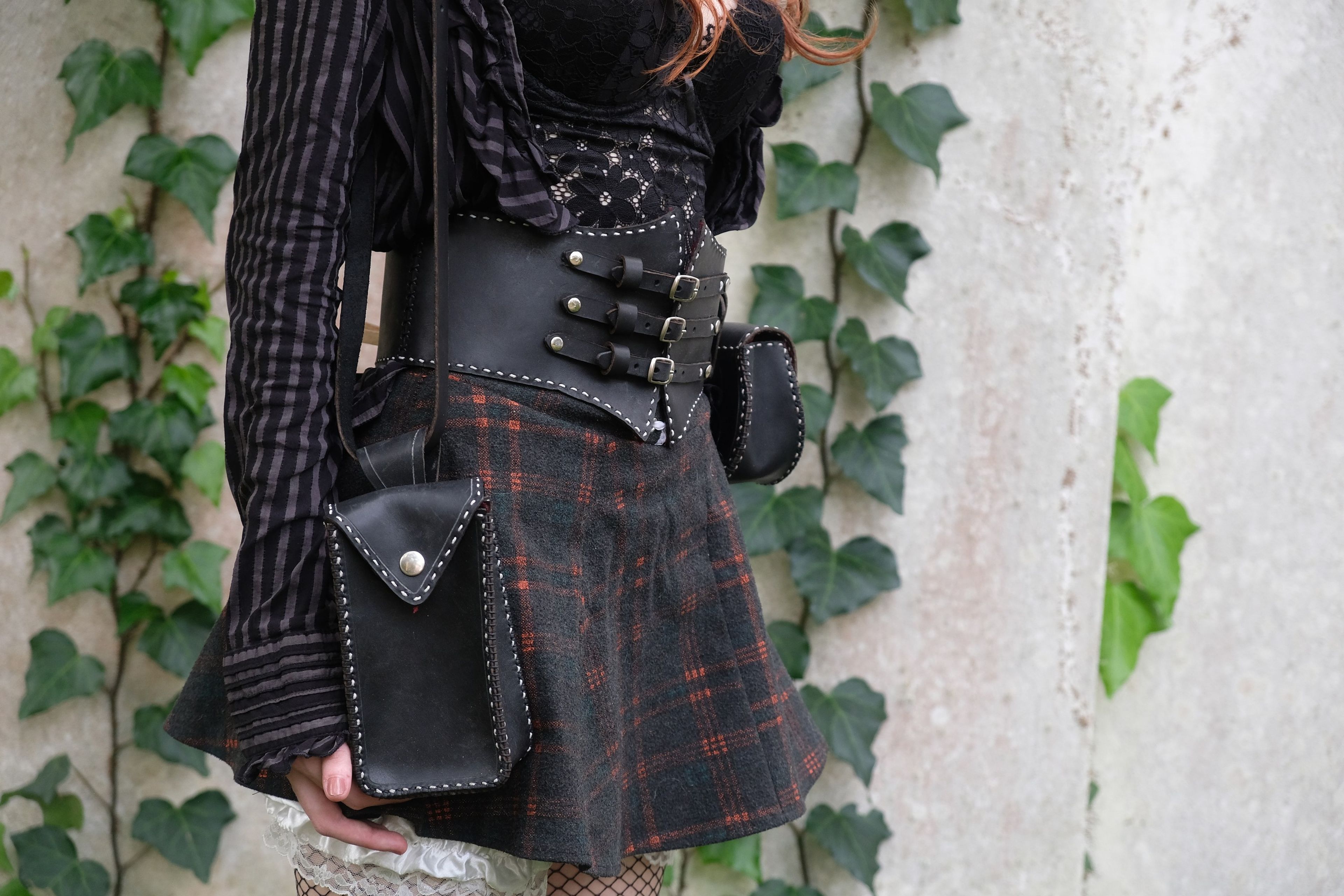 Black leather corset belt with buckles over a plaid skirt, with a black shoulder bag and pouch, against an ivy wall