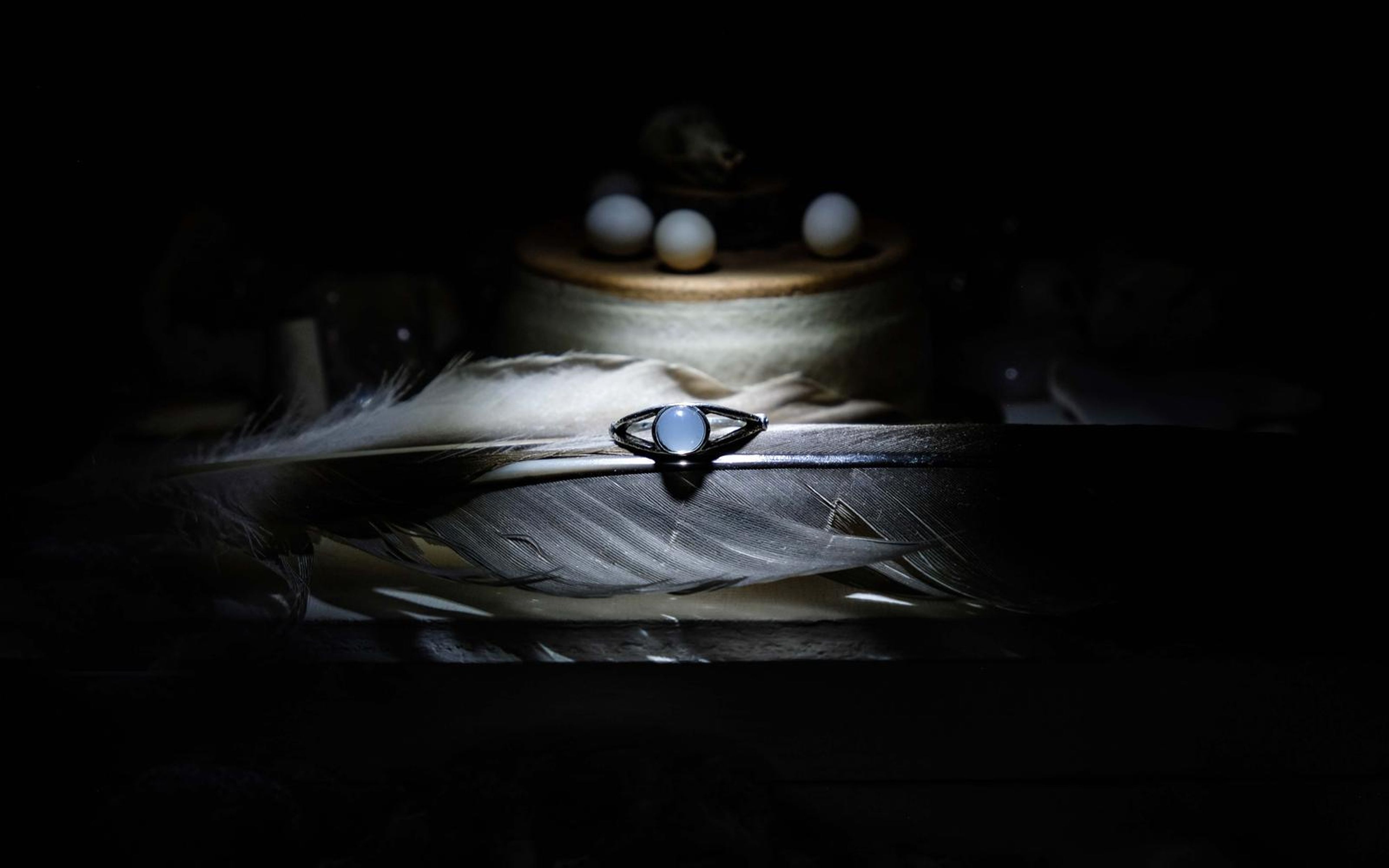 Silver ring with a pale blue stone resting on a feather, with a skull and spheres on a wooden stand above in dark light