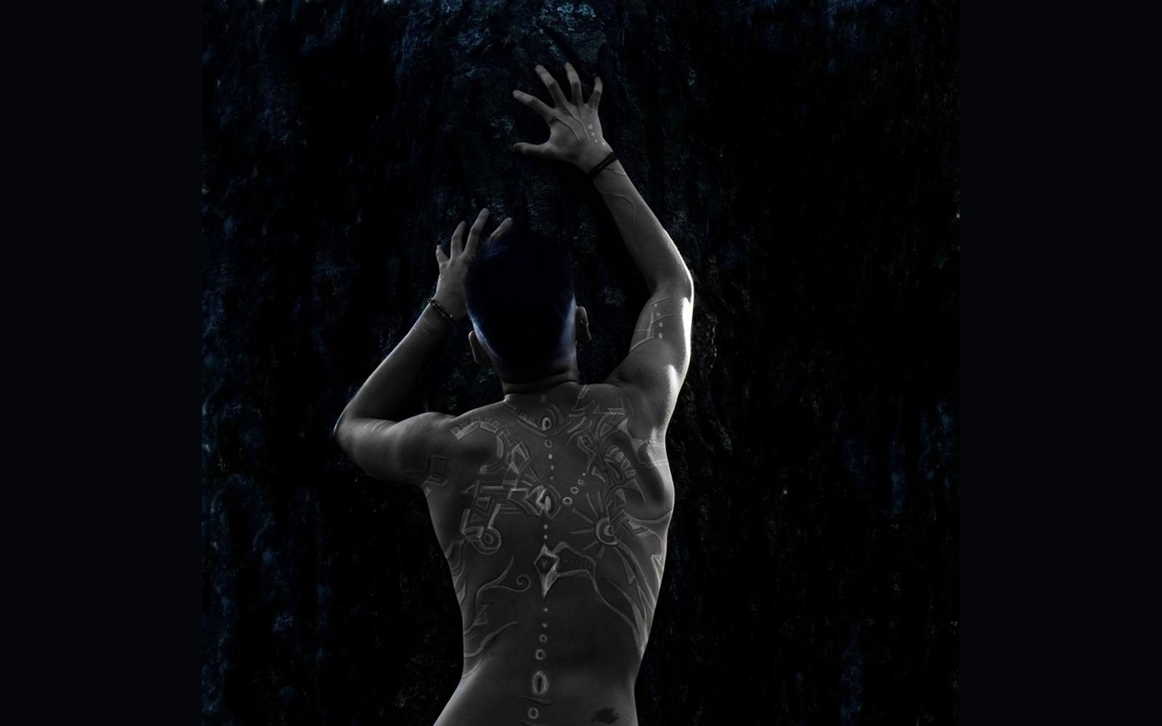Tighter crop of a figure from behind with painted back and raised hands, lit from the left on a dark wall
