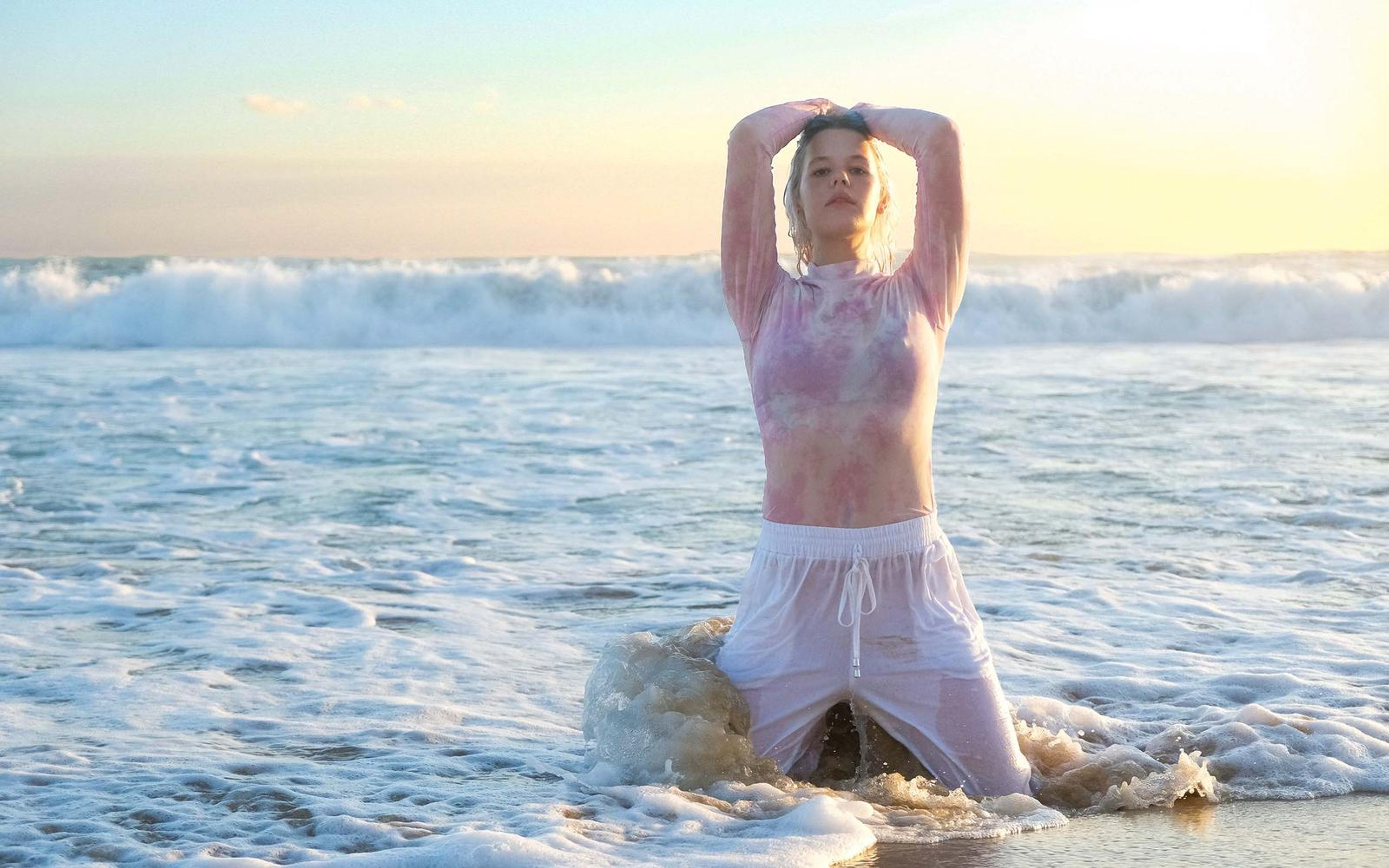 Wider view of a woman kneeling in surf with hands clasped above her head, foam breaking around her knees