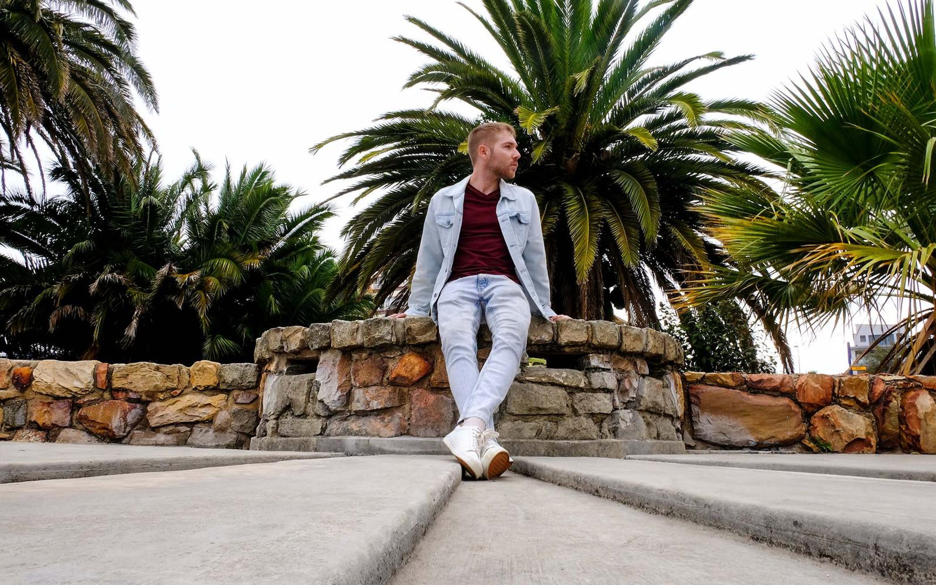 Man seated on a stone wall under palm trees, denim jacket, light jeans, white sneakers, low angle