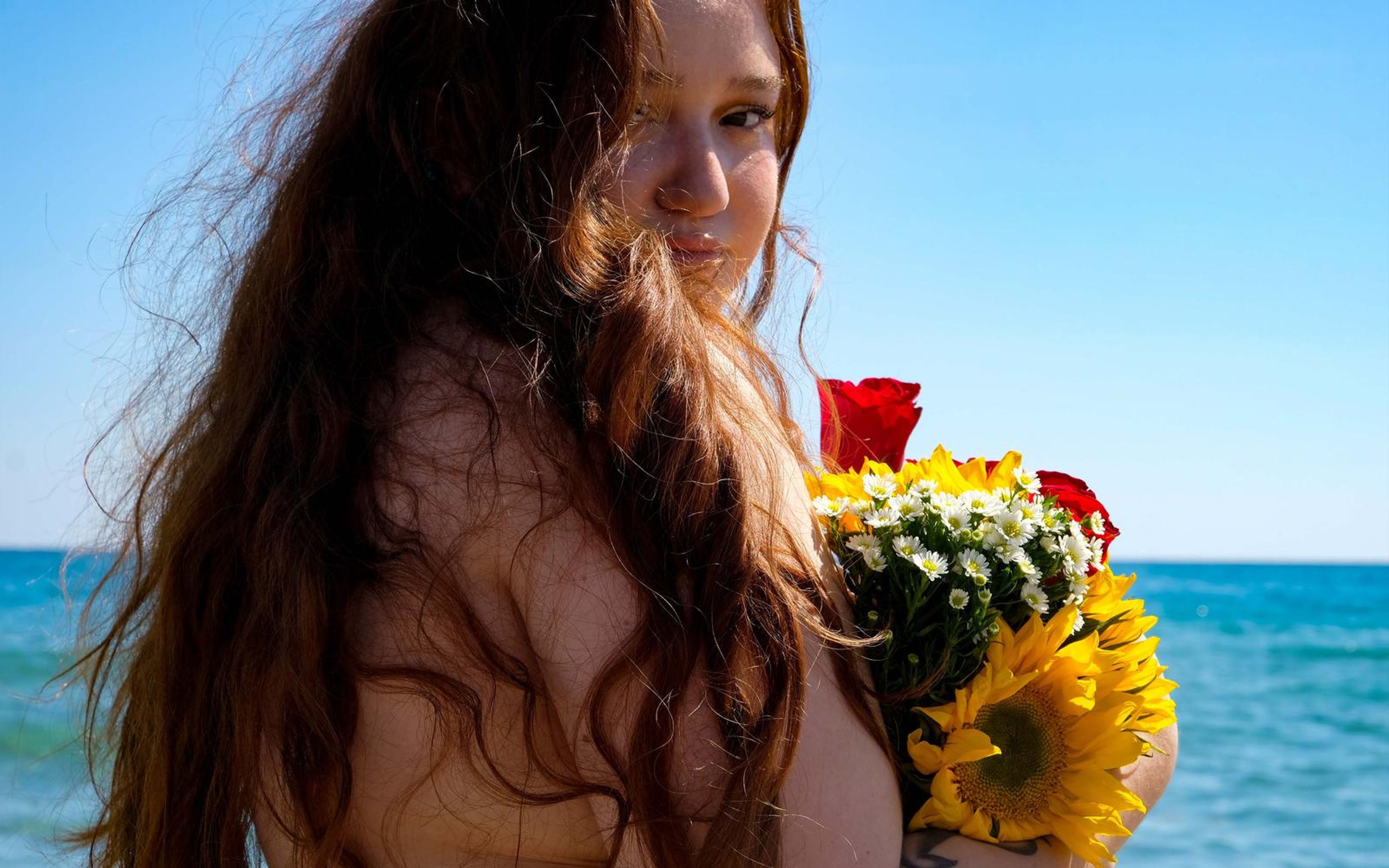 Close portrait of a woman with long windswept auburn hair across her face, a sunflower bouquet at her side and a clear blue sky and sea behind