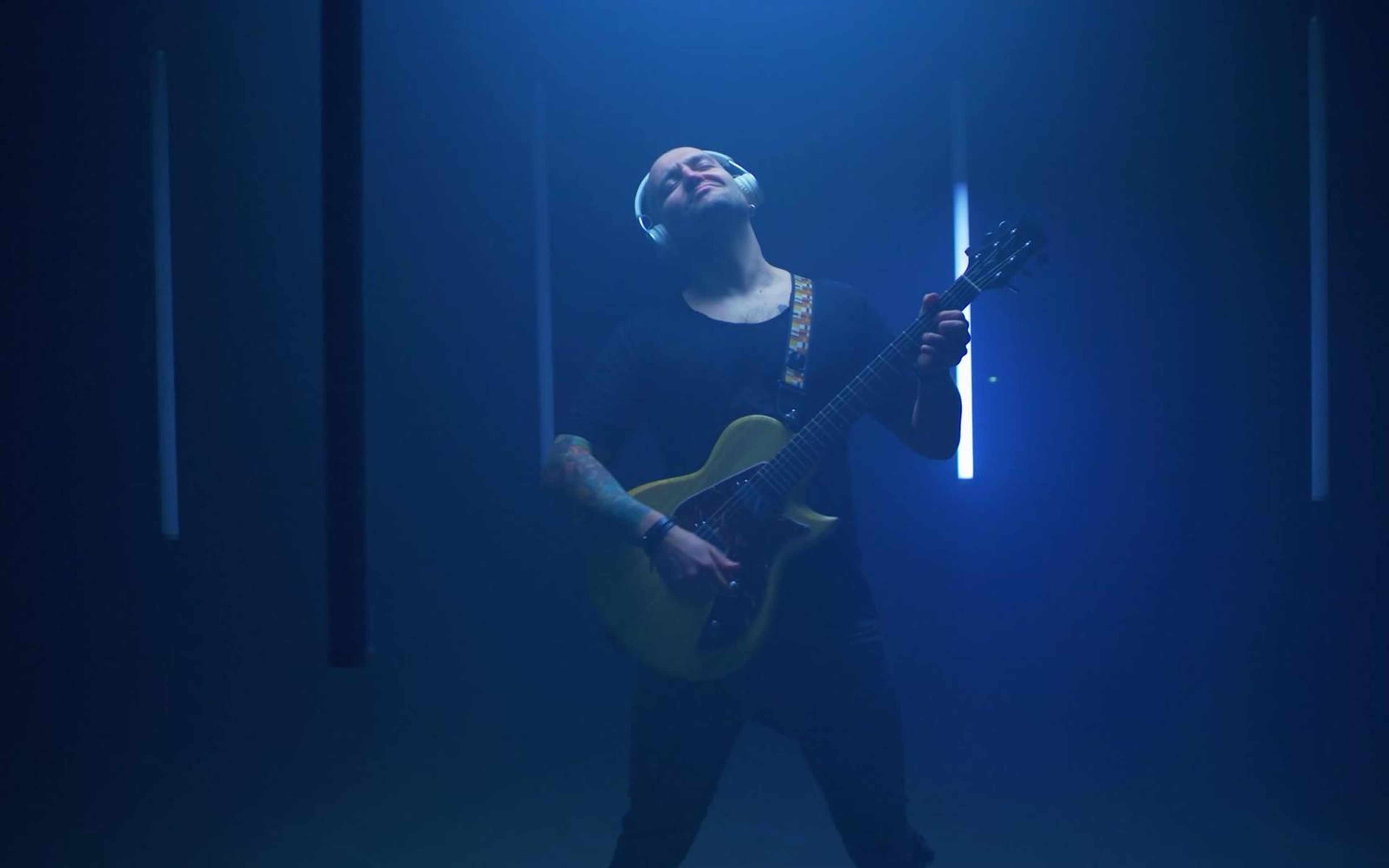 Guitarist in dark clothes and headphones playing in deep blue light between vertical light tubes