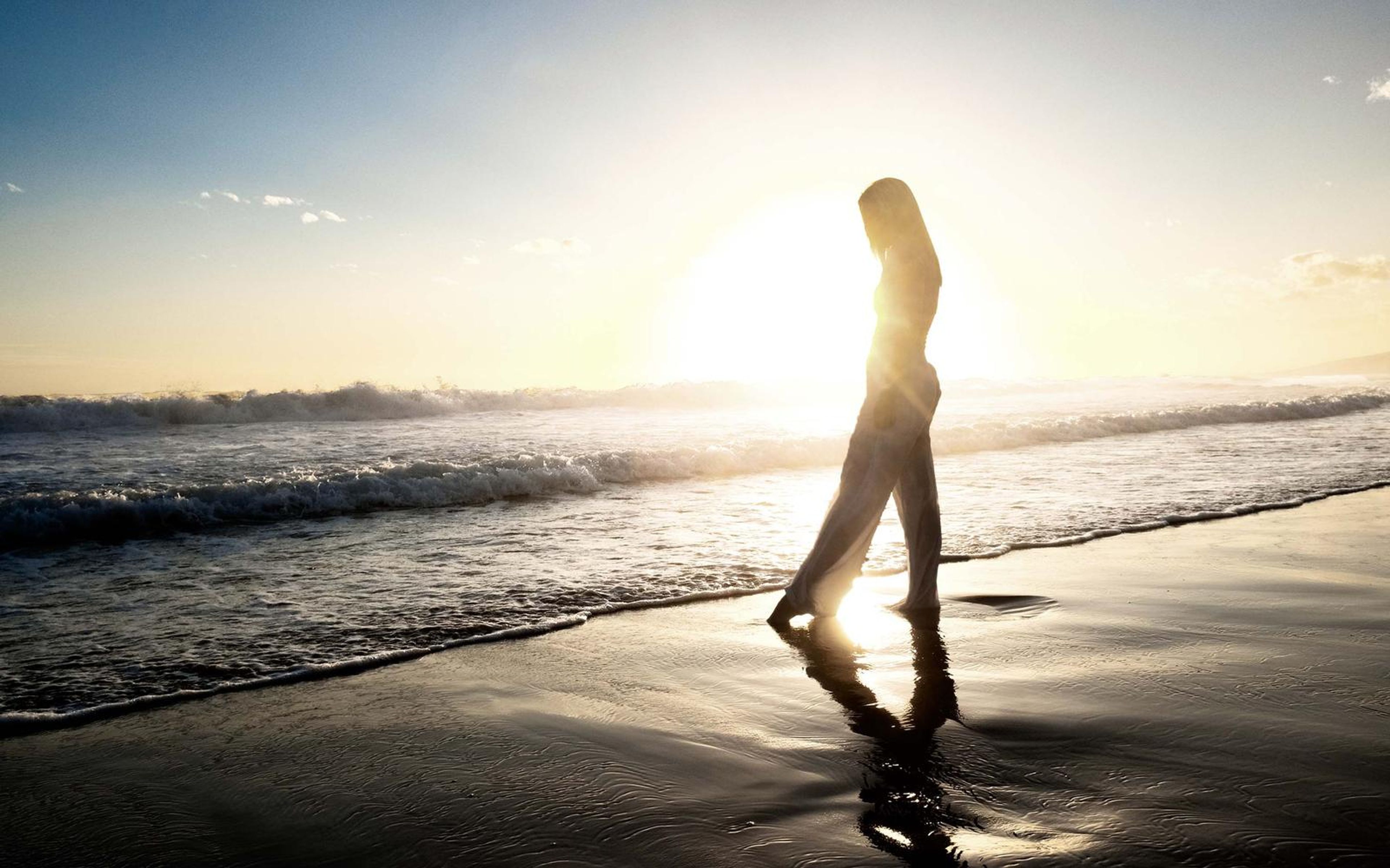 Woman silhouetted walking in the shallows with the sun behind, reflections on the wet sand