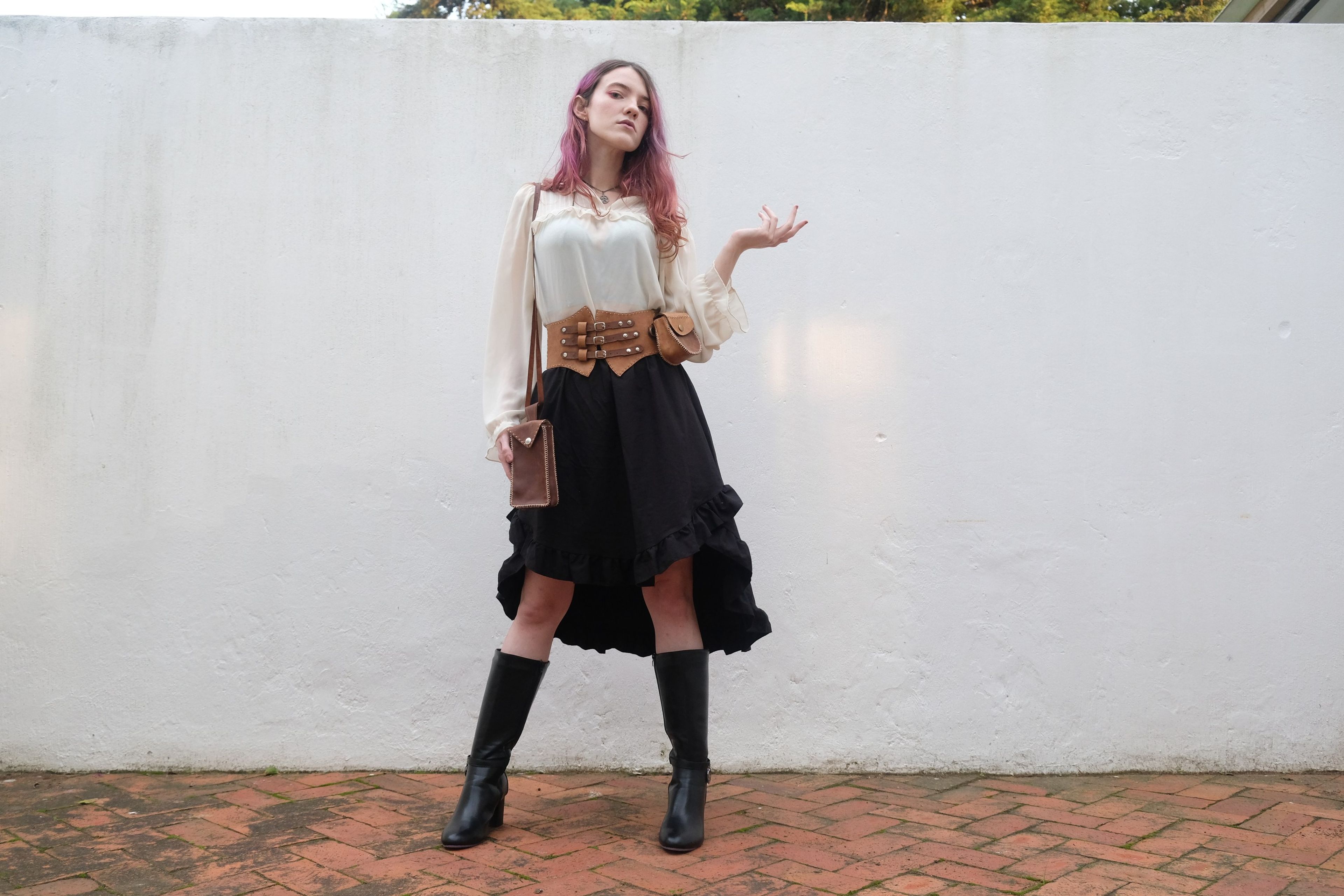 Woman standing on brick paving in boots, a ruffled skirt and a leather corset belt, one hand raised