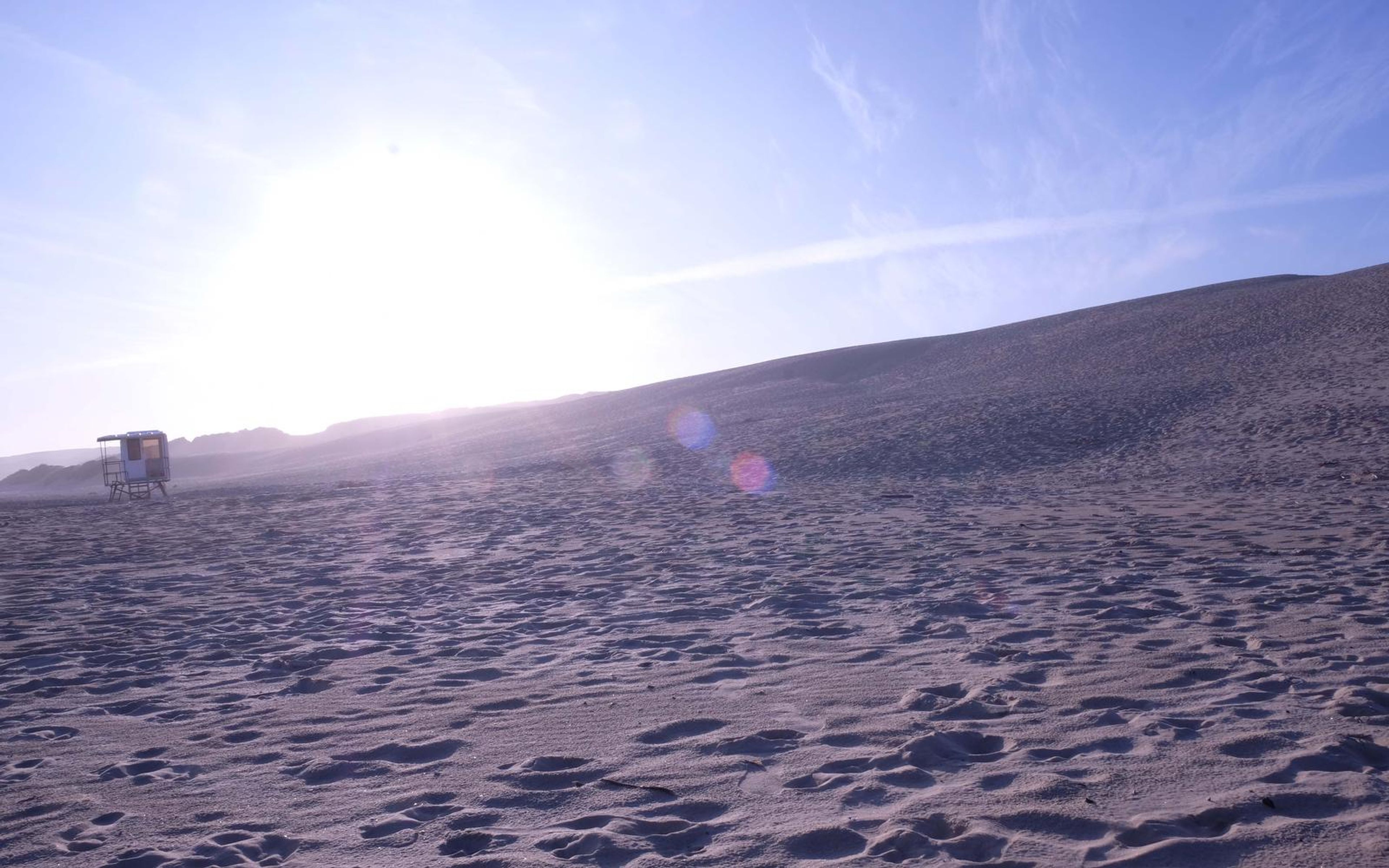 Low sun flaring over a dune slope with scattered footprints in the foreground sand