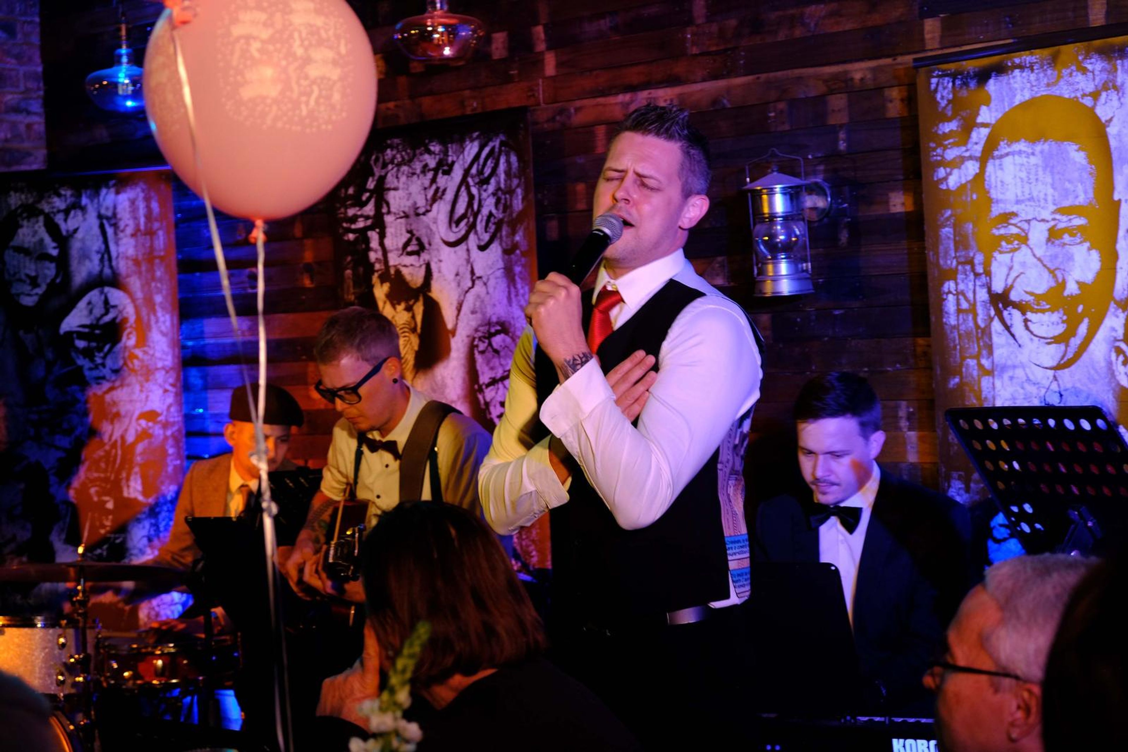 Singer in a waistcoat singing beside a balloon in front of a jazz mural