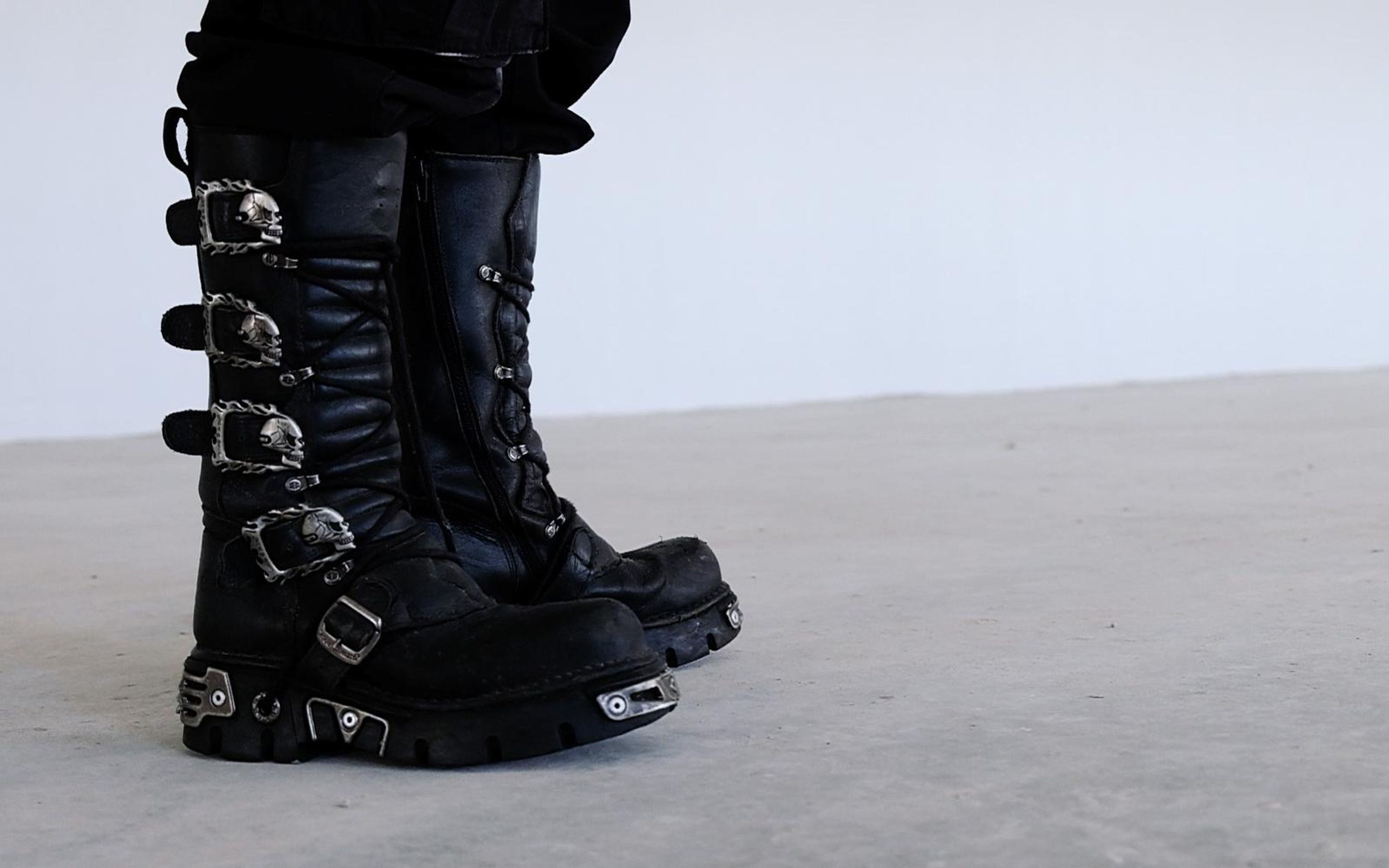 Rear three-quarter view of tall black boots with skull buckles standing on a pale concrete floor