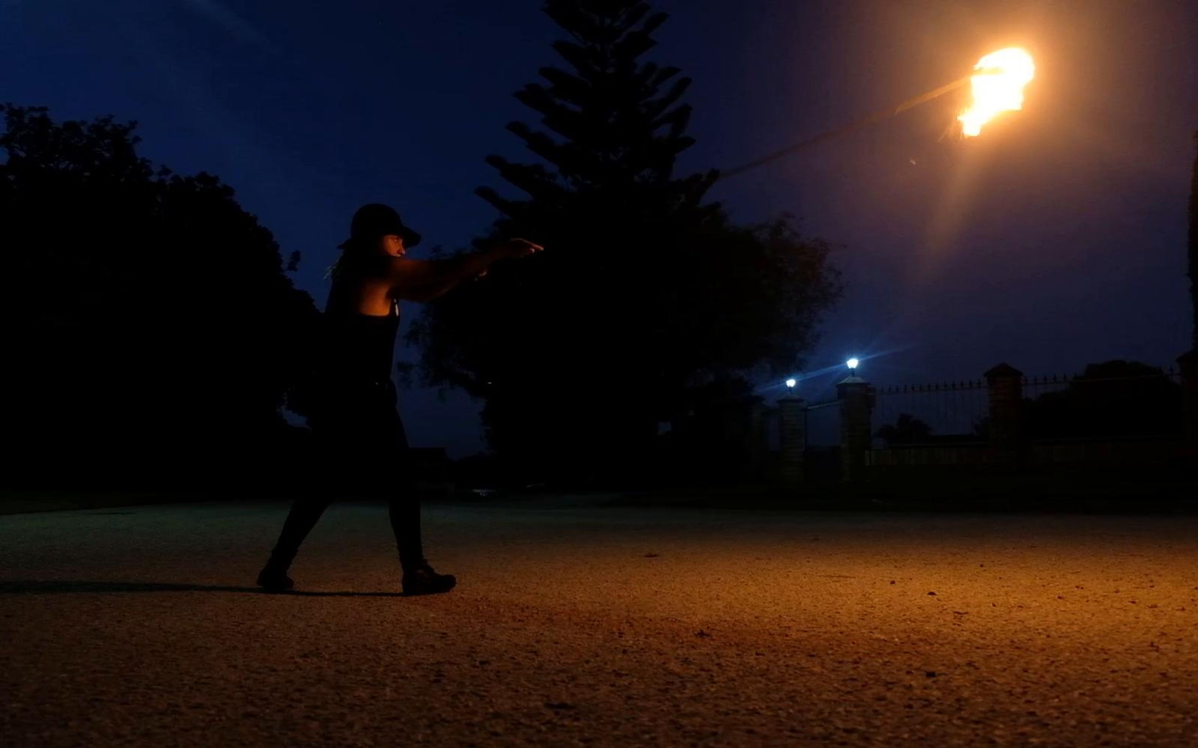 Two performers silhouetted in a car park with a flame held high at the right